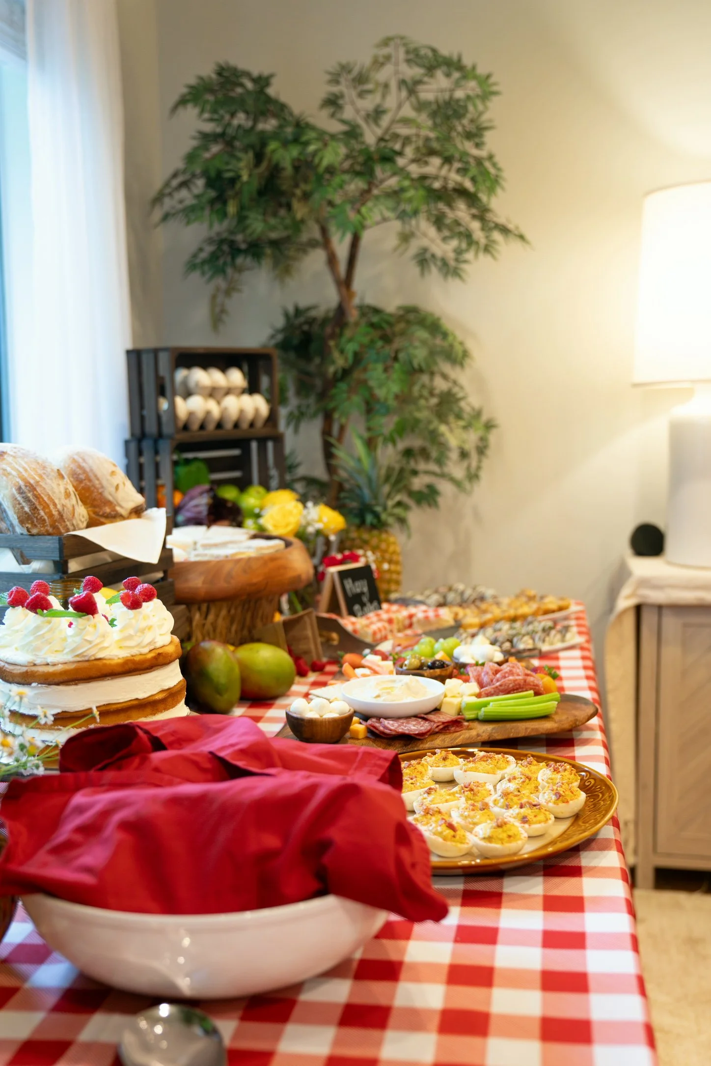 A table with a red and white checkered tablecloth set for a buffet or brunch, featuring a layered cake with whipped cream and raspberries, various cheeses, meats, sliced vegetables, deviled eggs, and baked dishes, in a room with natural light, a larg