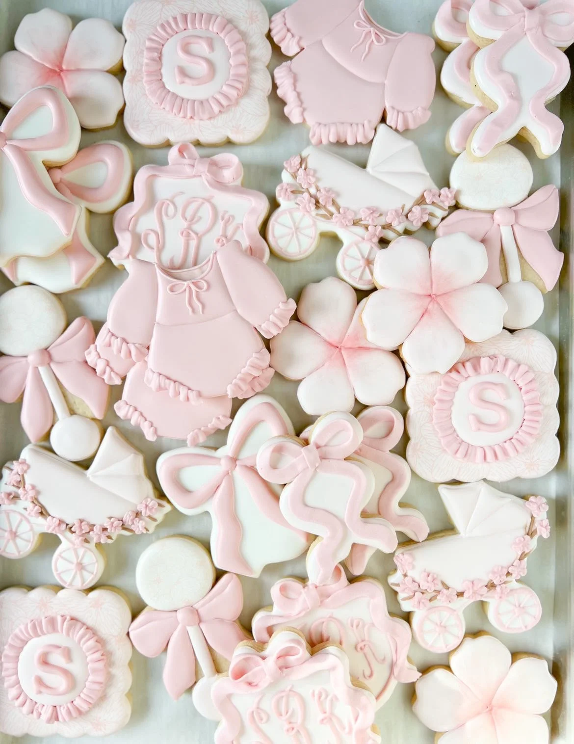 Collection of pink and white baby shower cookies, including flowers, baby clothes, baby stroller, bows, and monogrammed cookies with the letter 'S'.