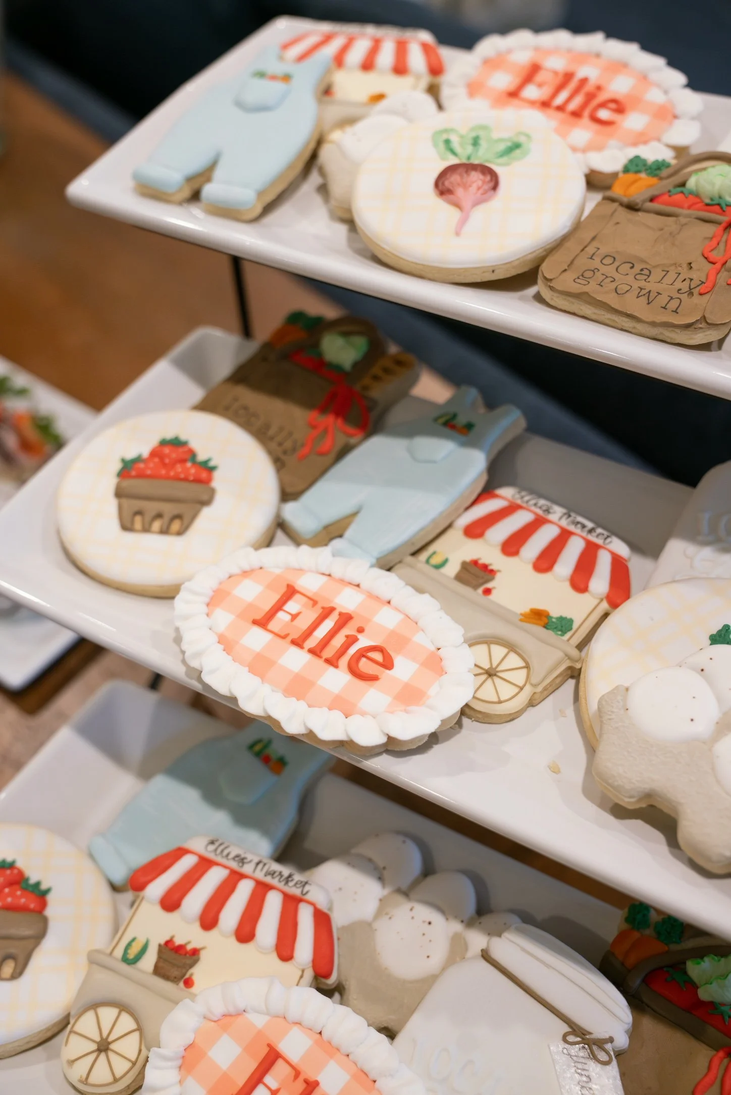Decorated cookies with farm and market themes, including a carrot, a wagon, and a sign that reads "Ummagood" on festive backgrounds, arranged on white trays.