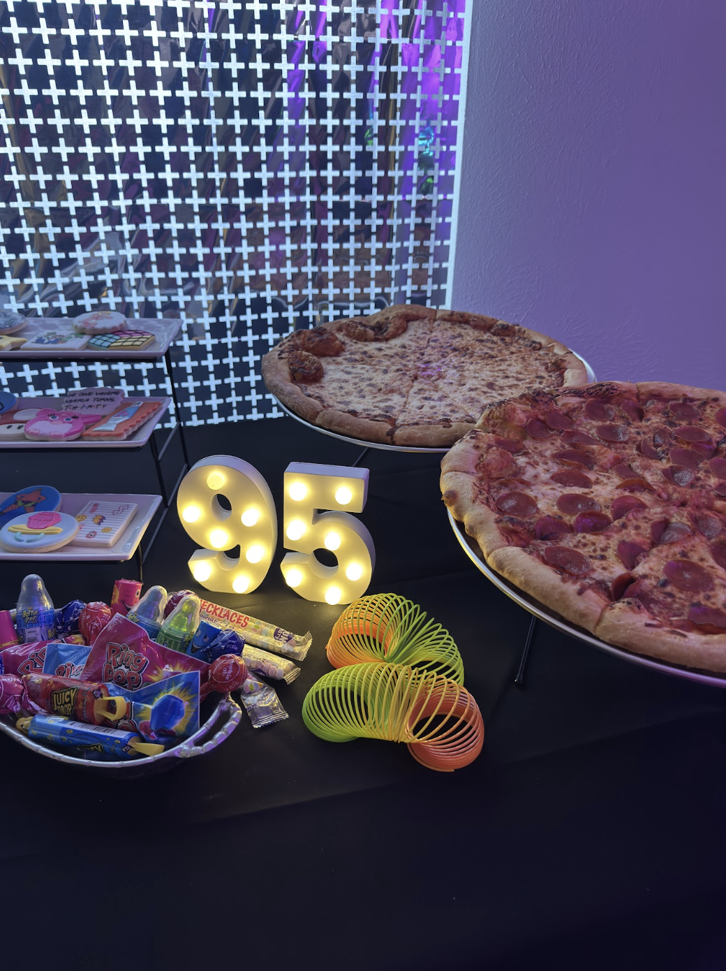 Table with two pizzas, a basket of assorted candies, colorful spiral toys, illuminated number '95', and a decorative background with purple lighting.