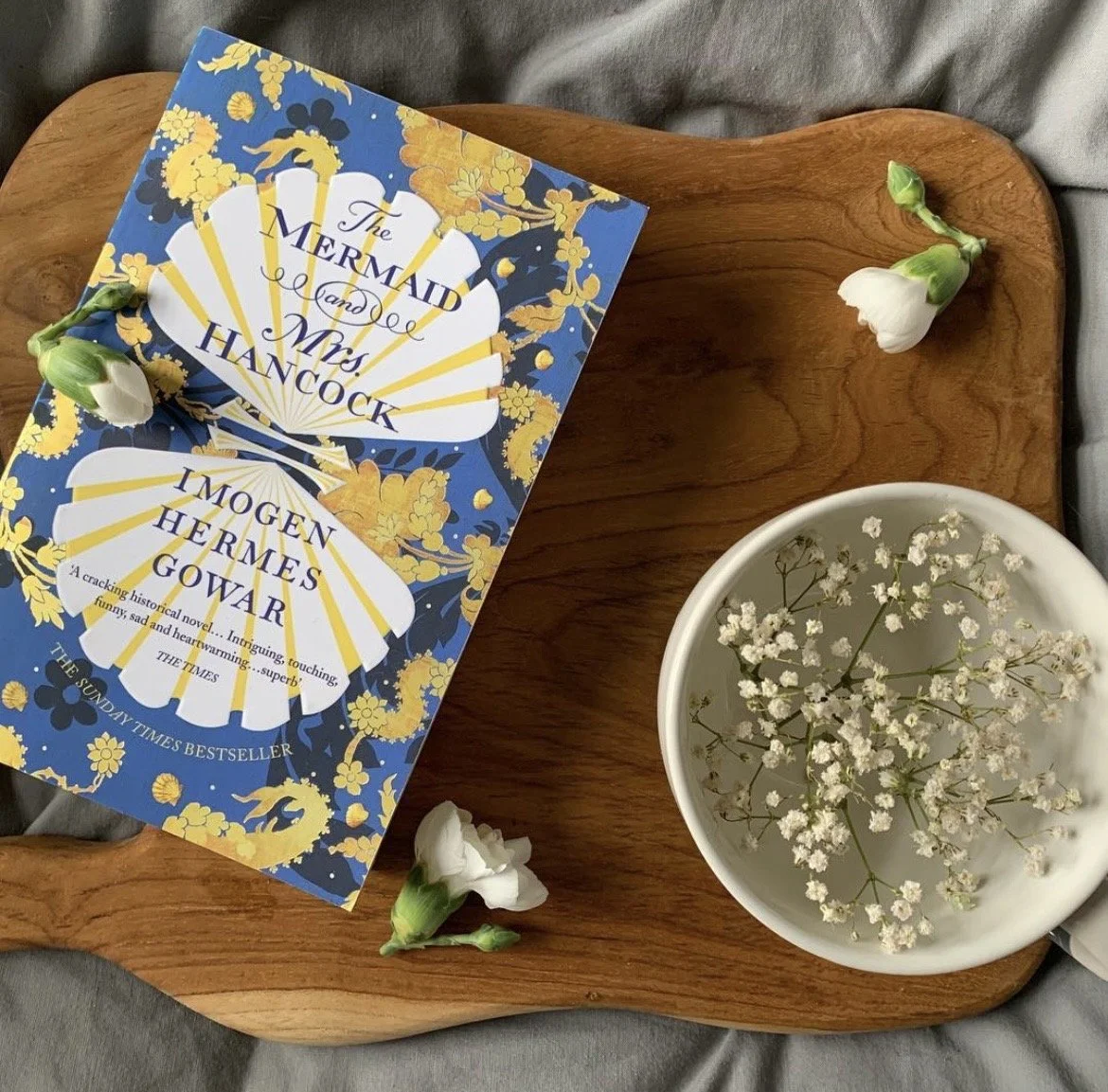 A wooden tray holding a copy of the book "The Mermaid and Mrs. Hancock" by Imogen Hermes Gower, three white flowers, and a white bowl filled with small white flowers or baby's breath.