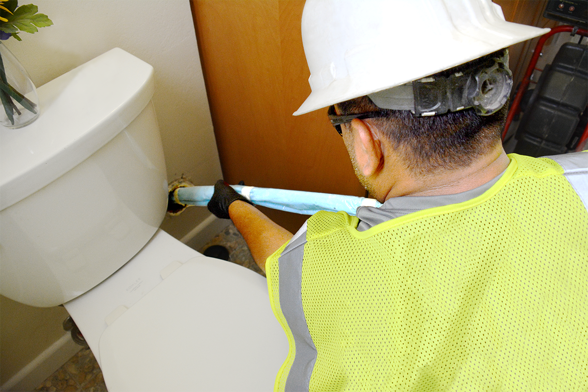 A worker wearing a white hard hat, yellow safety vest, and protective gloves is using nuflow technology to reline a pipe.