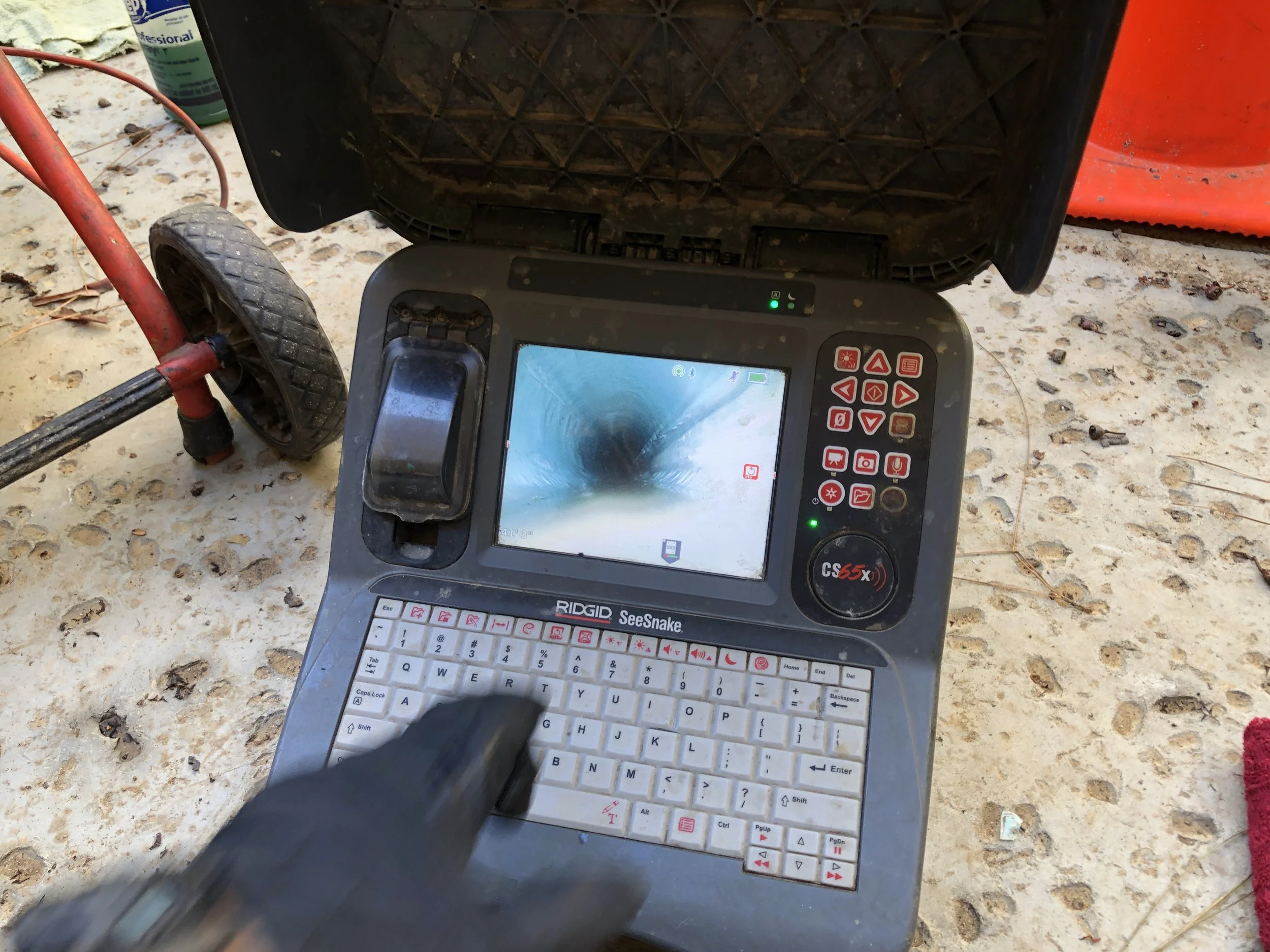 Close-up of worker visually inspecting a pipe with camera instrumentation on a gravel surface, with red equipment and cleaning supplies in the background.