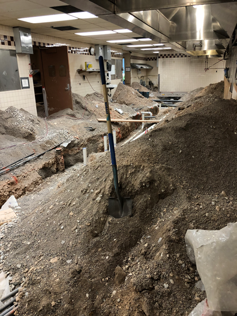 underground construction site with piles of dirt, a shovel stuck in the dirt, and pipes for plumbing or electrical work in a room with a tiled ceiling and various equipment.