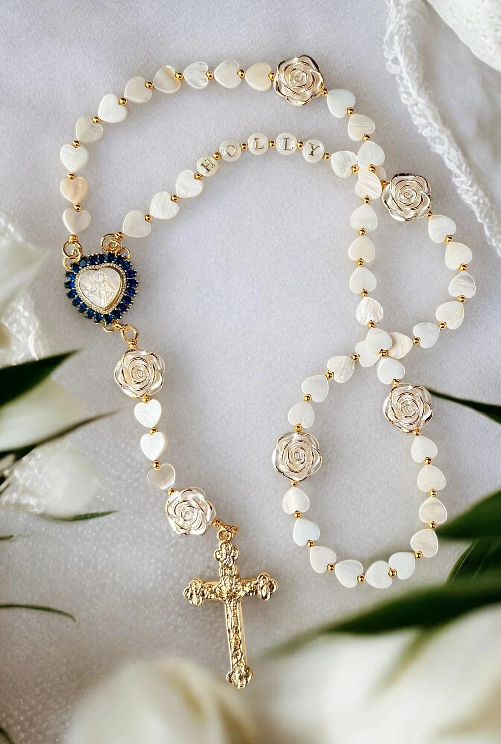 A white rosary with gold accents, featuring gold rose-shaped beads and a gold crucifix, with the words 'Holy' spelled out in silver letter beads.