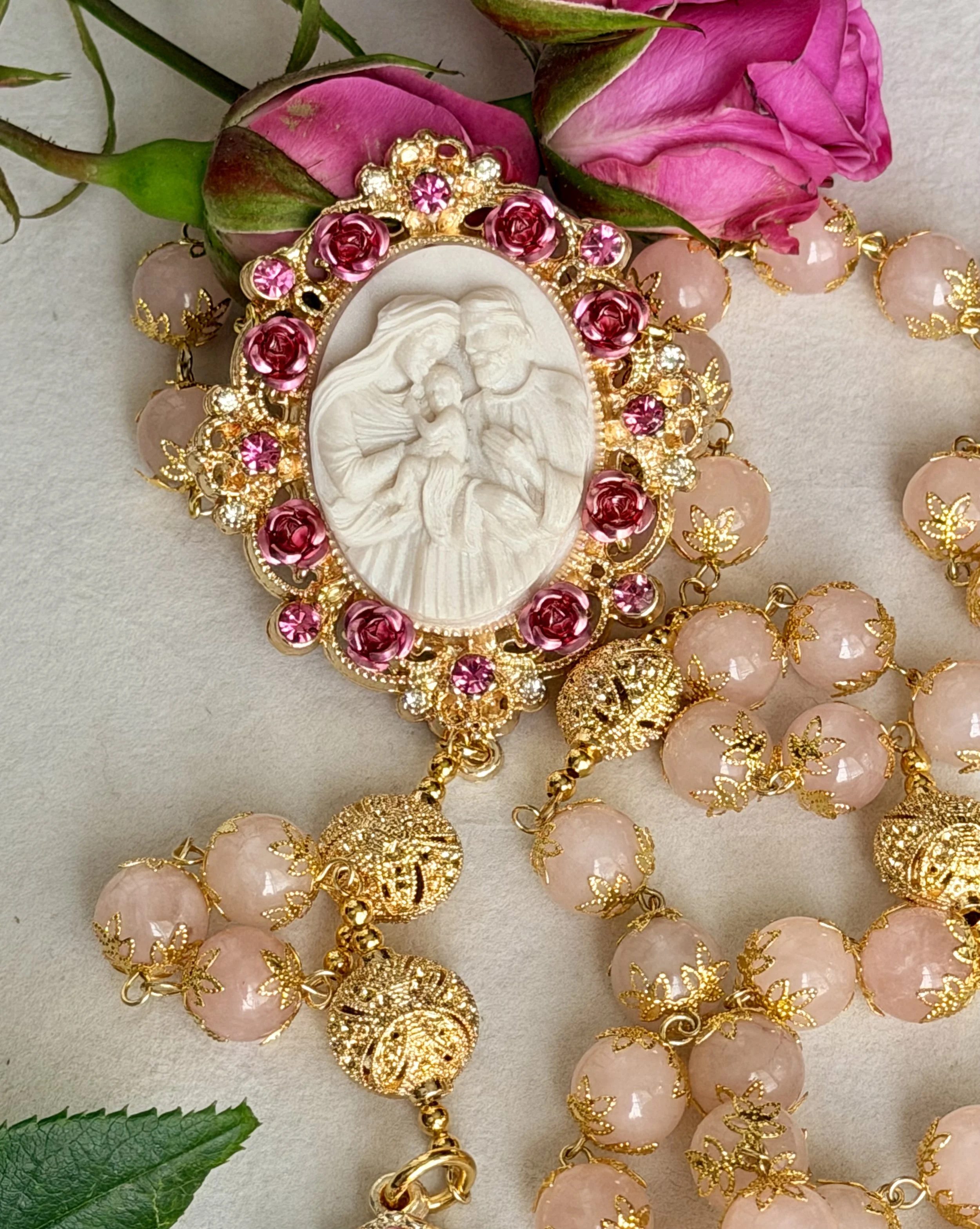 A decorative religious item with a carved white relief of Jesus and Mary, surrounded by pink and gold embellishments, pink roses, pink beads with gold accents, and pink flowers, arranged with pink roses and green leaves in the background.