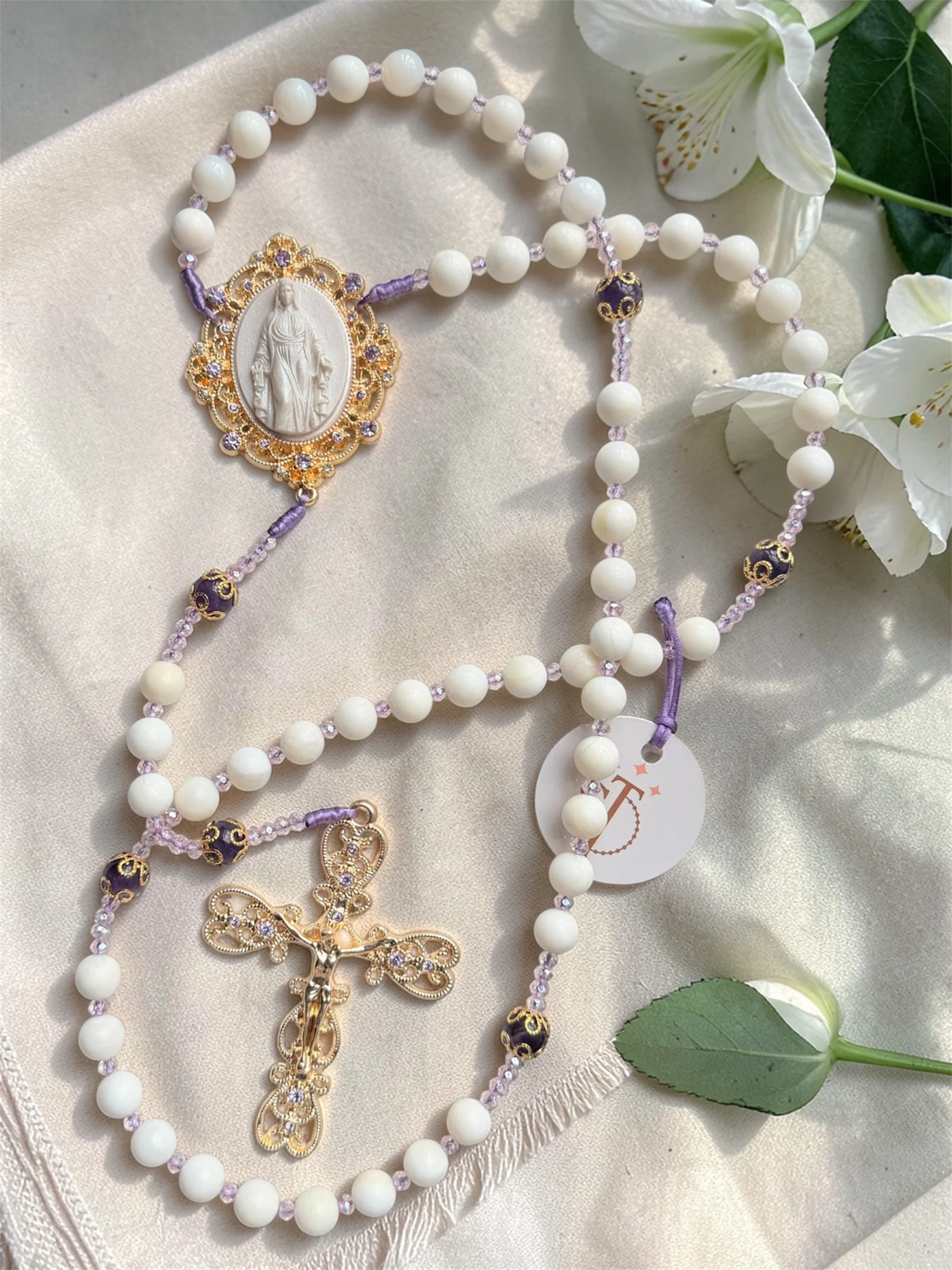 A close-up of a rosary with white beads, purple accents, and gold-colored religious medals and a crucifix, placed on a white fabric with white lilies nearby.