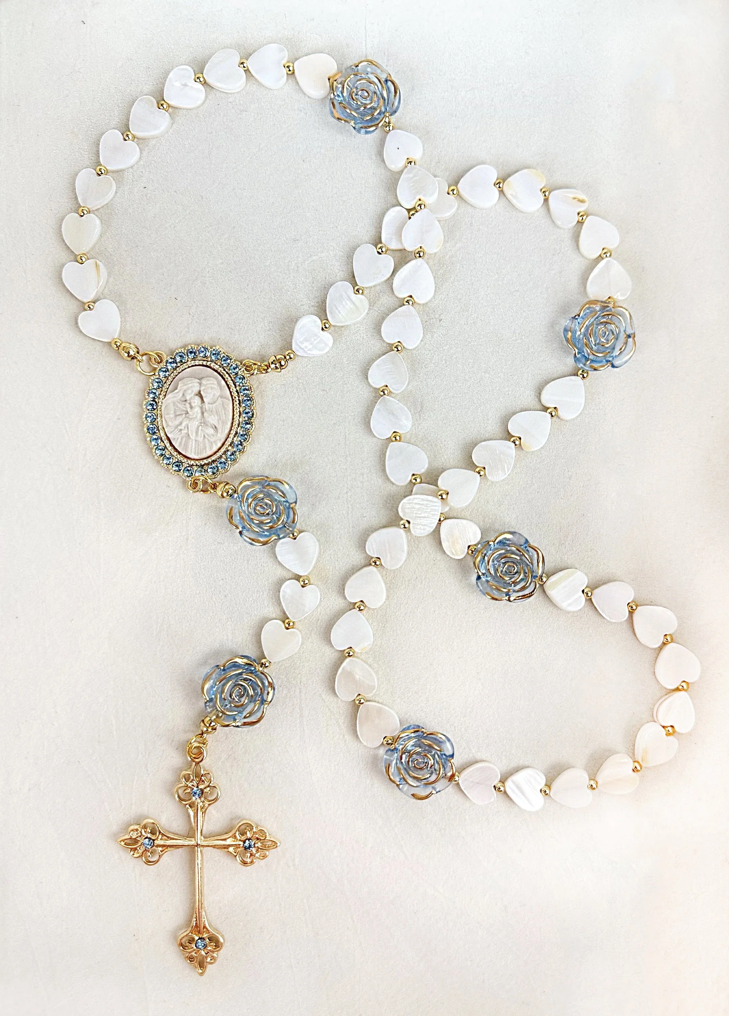 A rosary with white heart-shaped beads, gold links, blue and gold rose motifs, a circular medallion with a religious image surrounded by blue rhinestones, and a gold cross pendant encrusted with blue rhinestones.