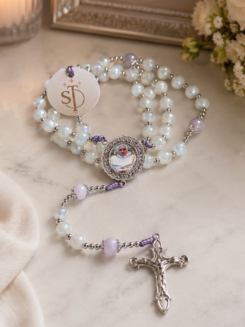 A rosary with white pearls and purple beads, featuring a photo of a person in the center, lying on a white surface next to a beige fabric, with a bouquet of white flowers in the background.