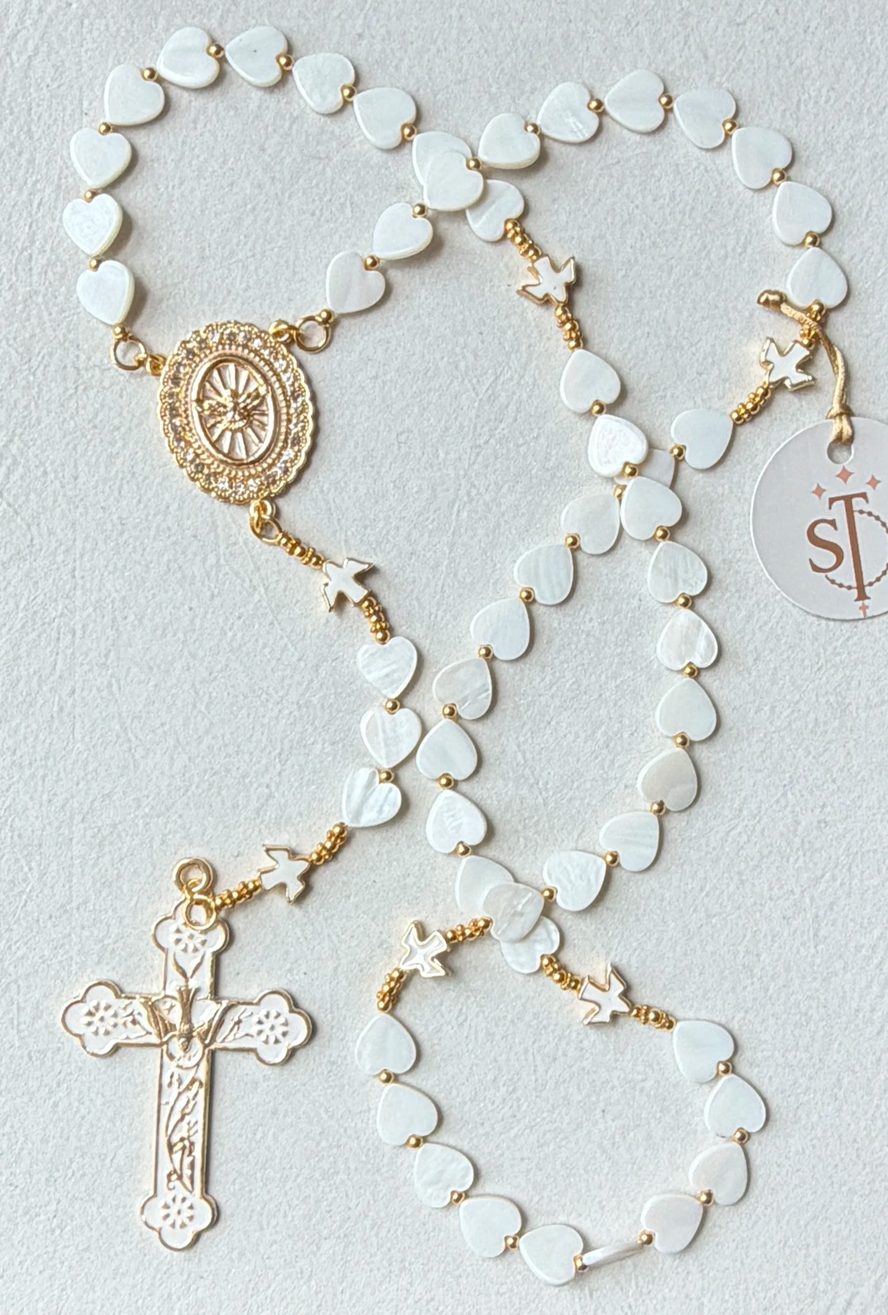 A white rosary with gold accents, featuring heart-shaped beads, a crucifix, and a medallion with a design.