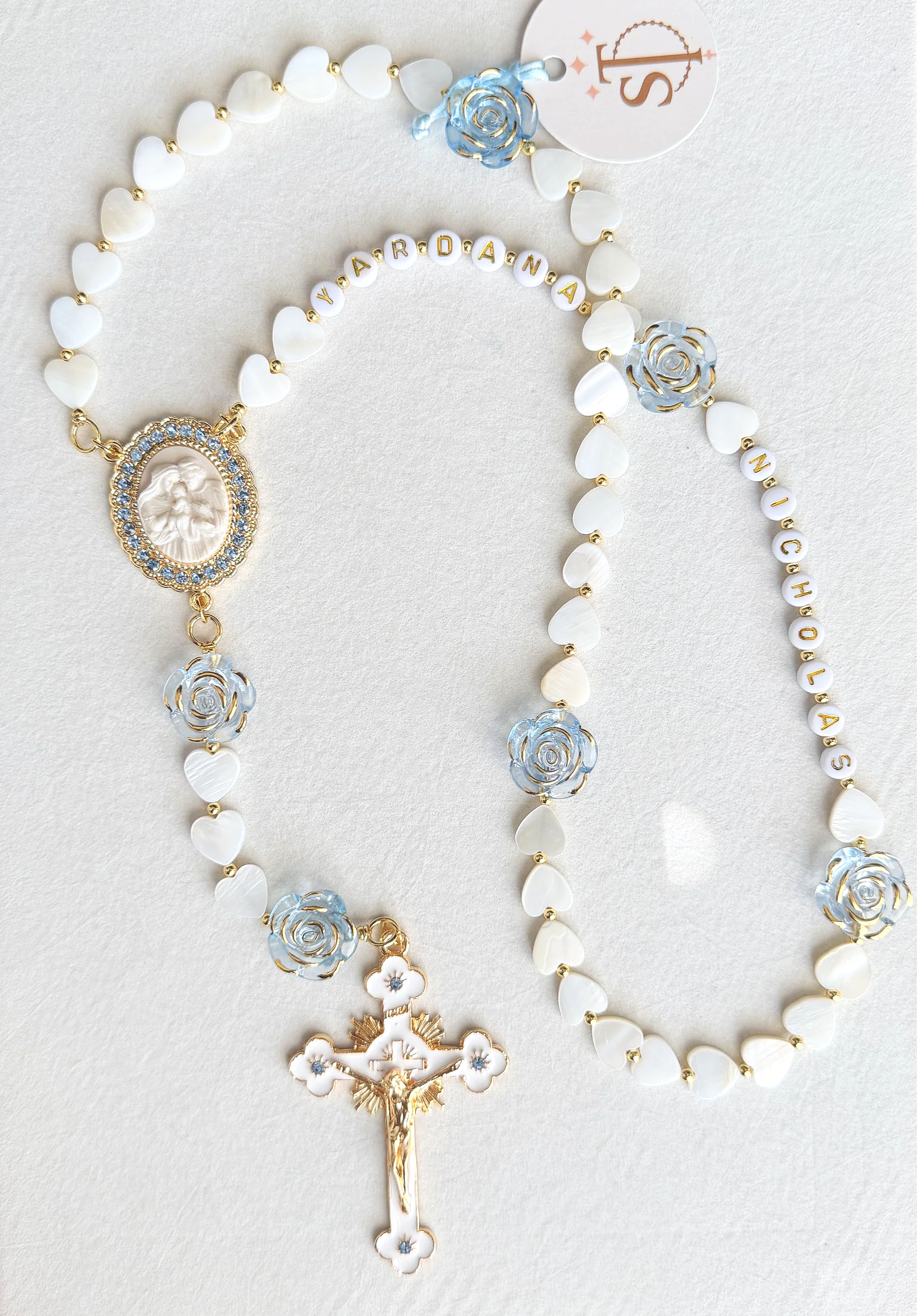 Close-up of a religious rosary with white heart-shaped beads, light blue and white floral accents, gold details, a cameo medallion, and a crucifix, laid on a light-colored surface.