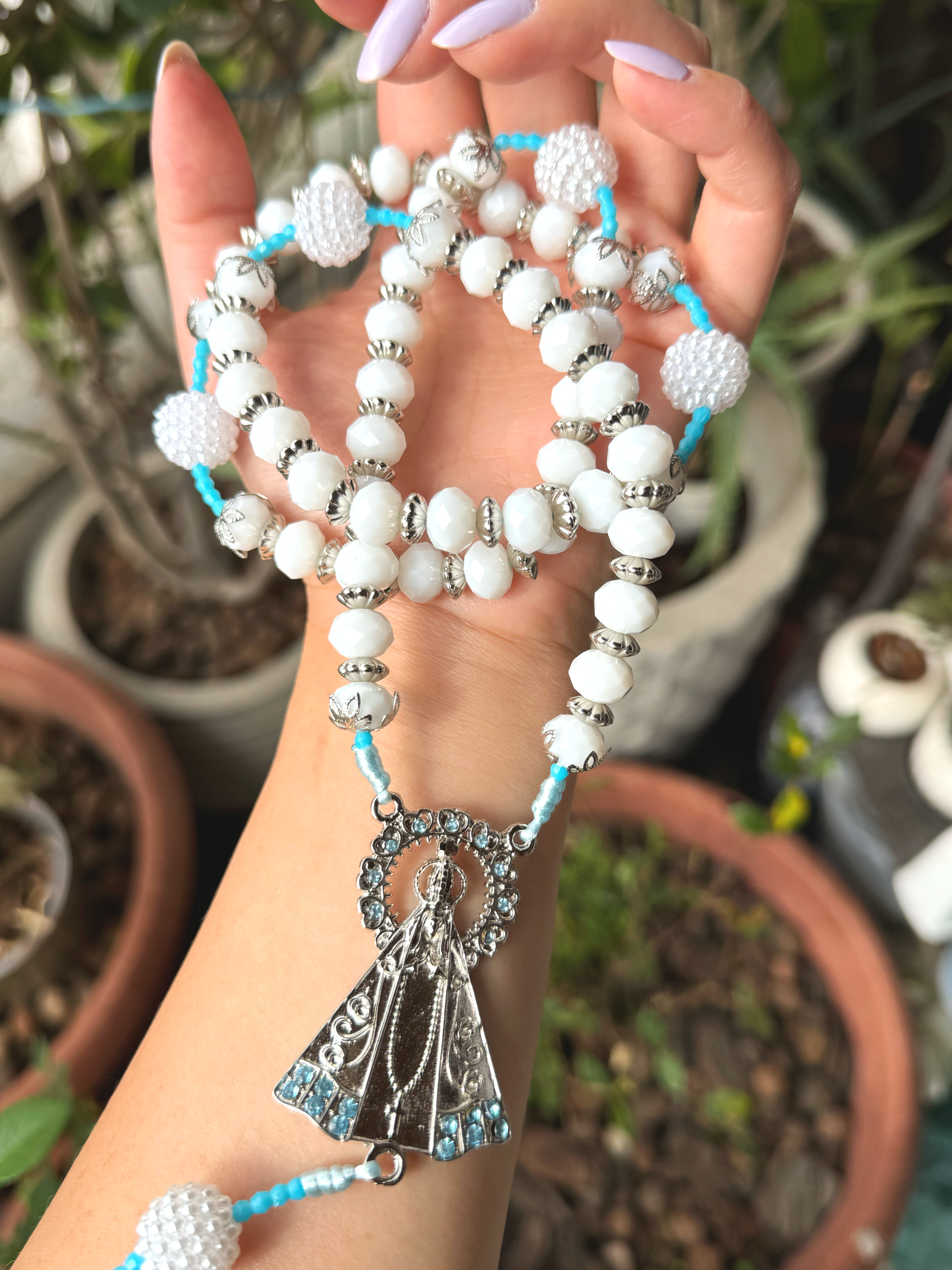 Close-up of a hand holding a silver and white beaded necklace with a silver angel pendant, with potted plants in the background.