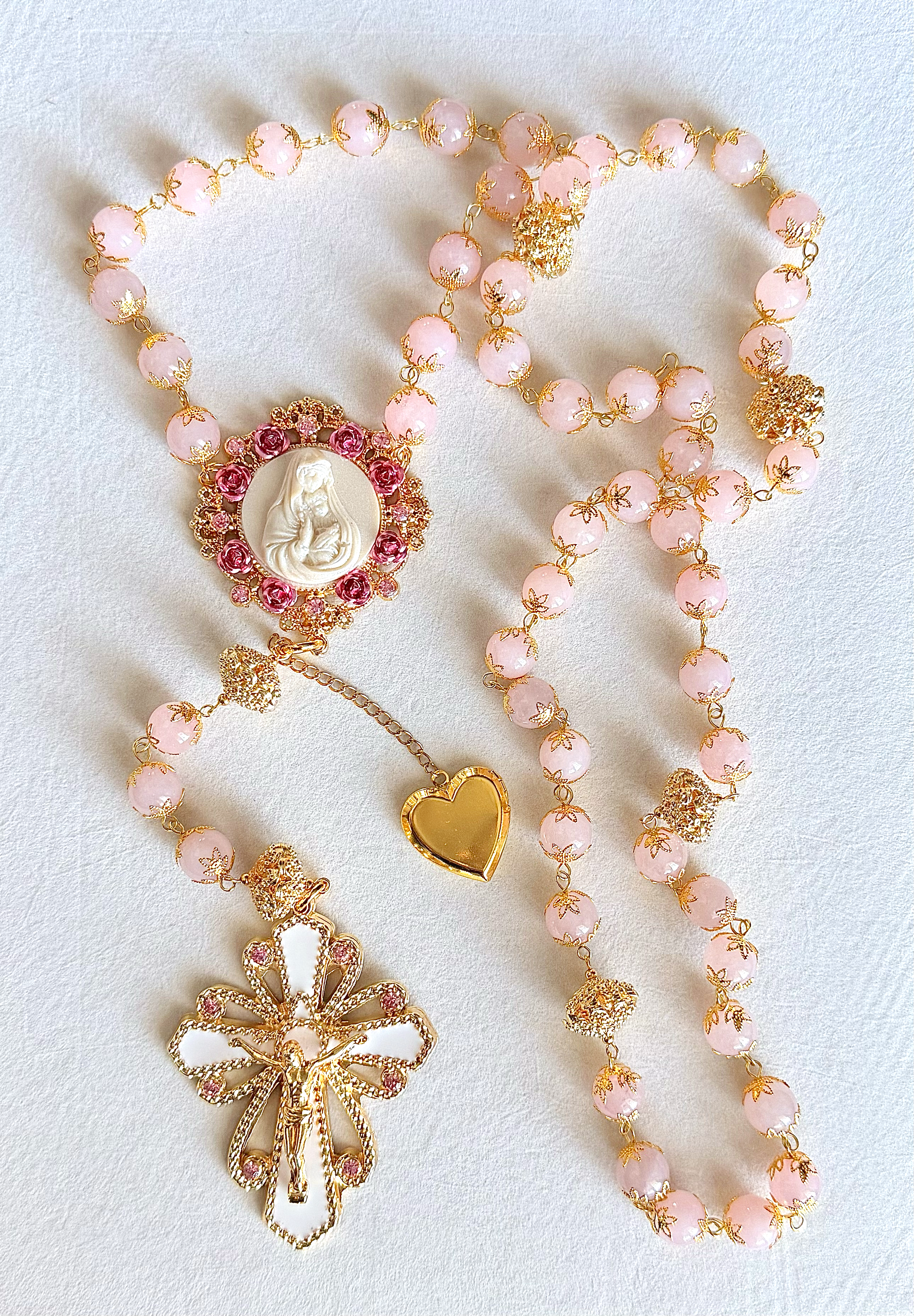 Collection of pink and gold jewelry including necklaces with floral and religious pendants, a gold heart charm, and a decorative bow pendant, arranged on a white fabric background.
