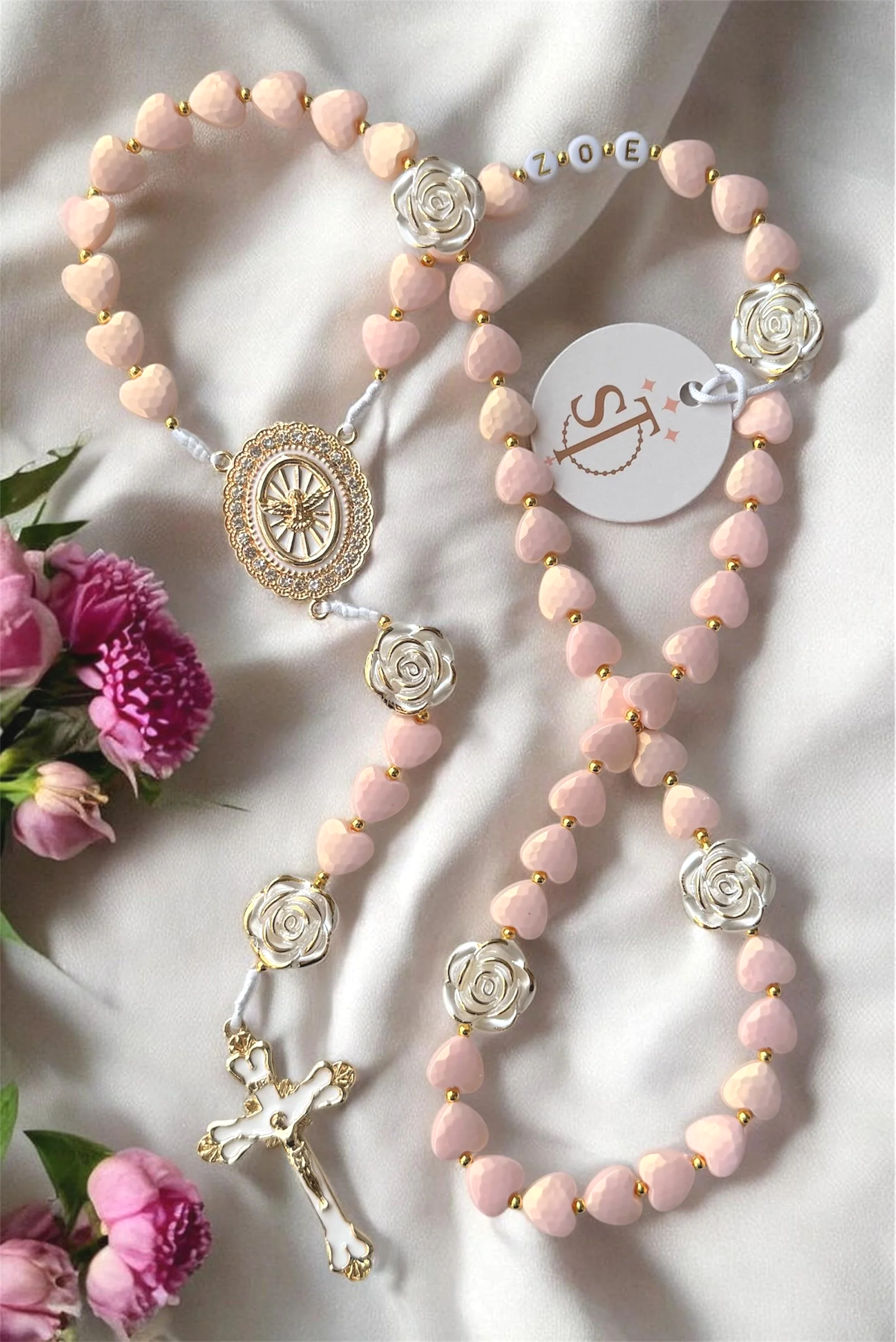 A pink prayer rosary with gold accents, featuring heart-shaped beads, silver rose charms, a decorative medallion with an angel, a gold cross, and an attached tag with a logo, placed on a cream satin fabric, next to pink flowers.