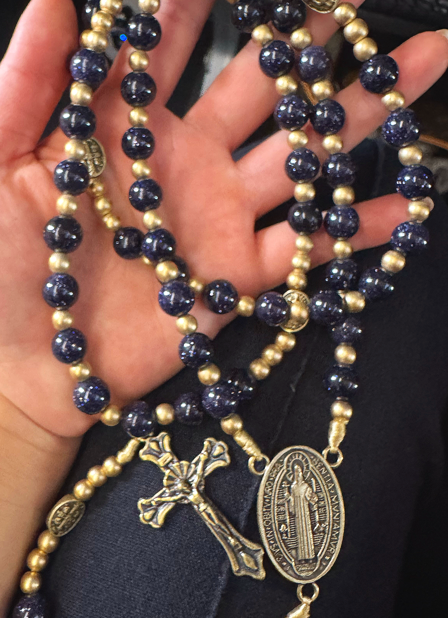 Close-up of a person's hand holding multiple religious and decorative necklaces, including gold and dark blue bead strands, with crosses and medallions.