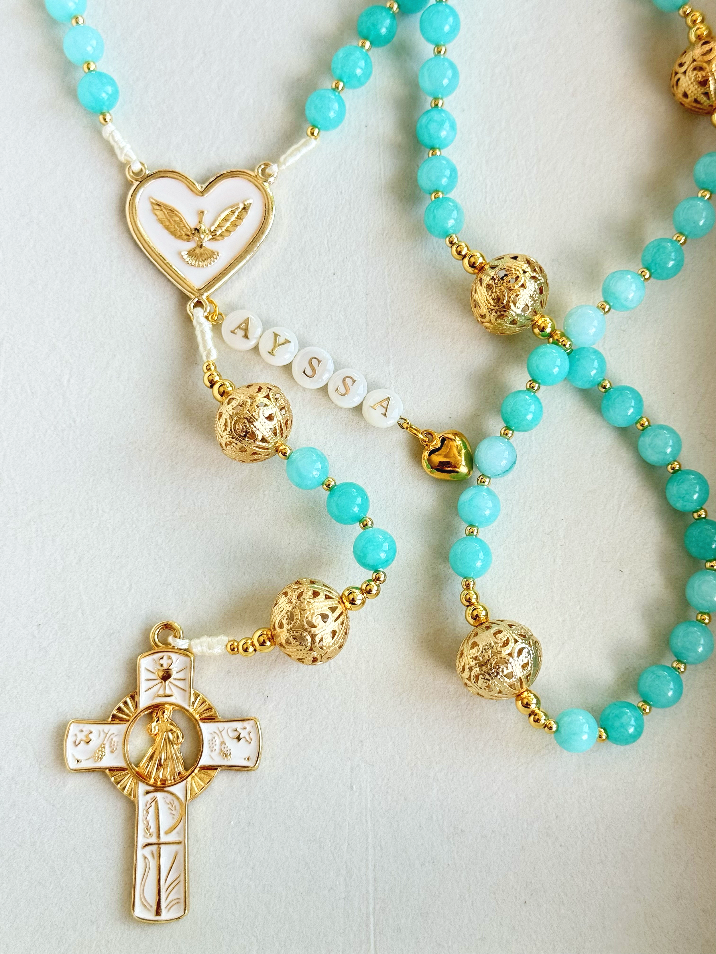 Close-up of turquoise beaded necklaces with gold beads and religious pendants, including a white and gold crucifix and a white and gold medallion with a religious figure.