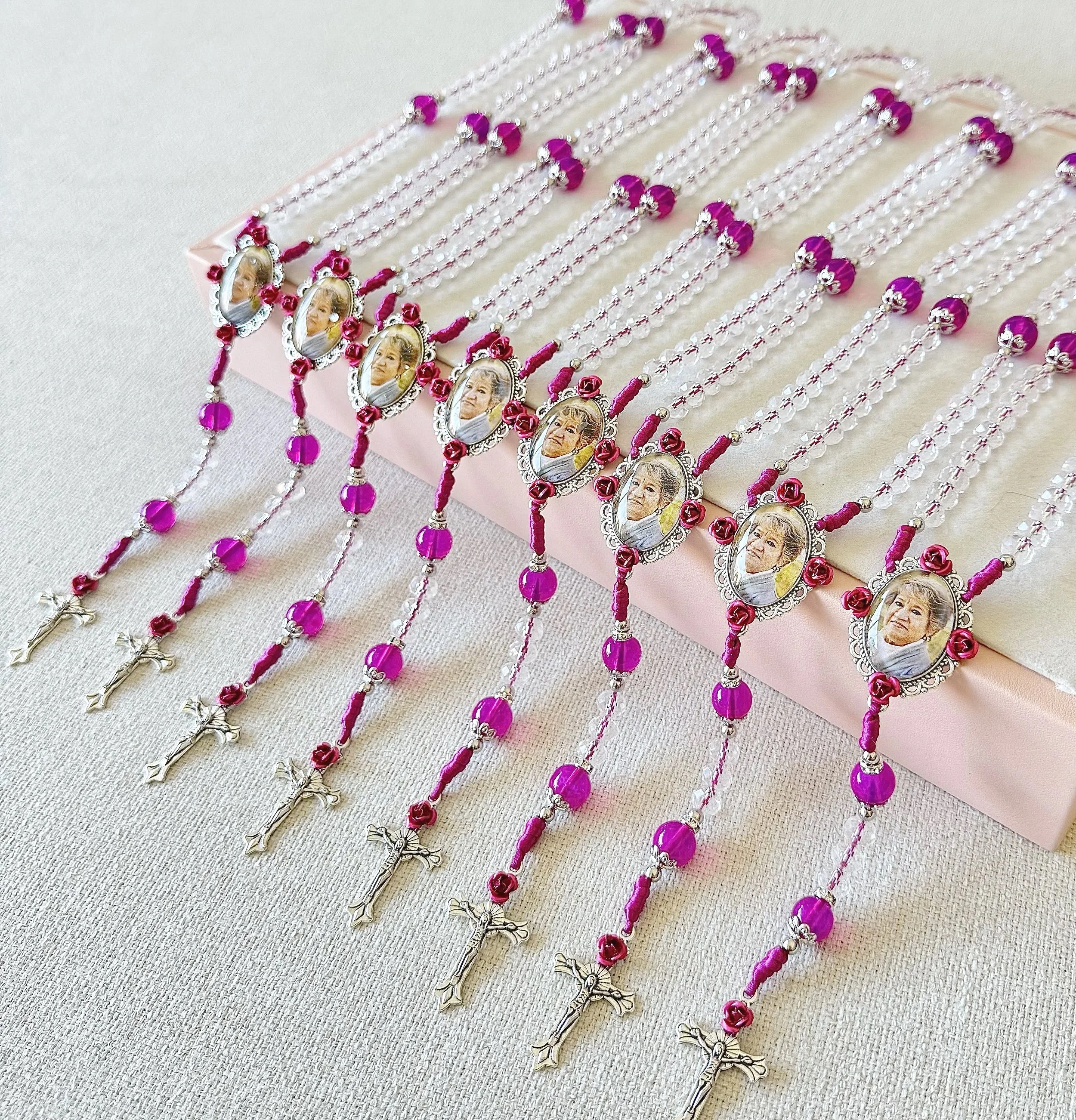 A display of rosary necklaces arranged in multiple rows. The necklaces feature purple beads, silver chain links, and photos of an elderly woman framed in decorative silver settings. Some necklaces have a silver crucifix at the end.