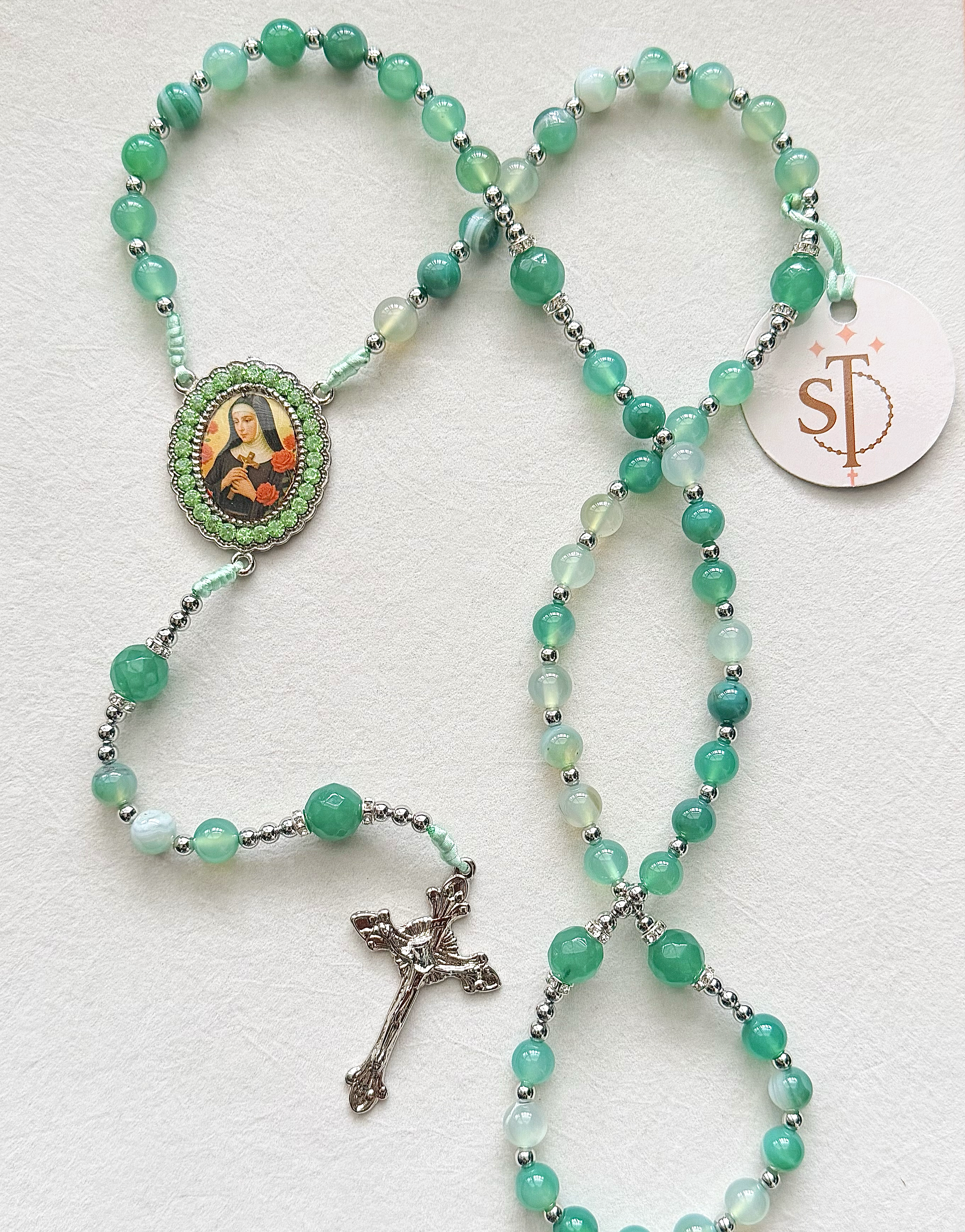 Multiple turquoise and white beaded rosary necklaces with silver accents, featuring a depiction of a saint, and an attached silver crucifix, laid on a white surface.