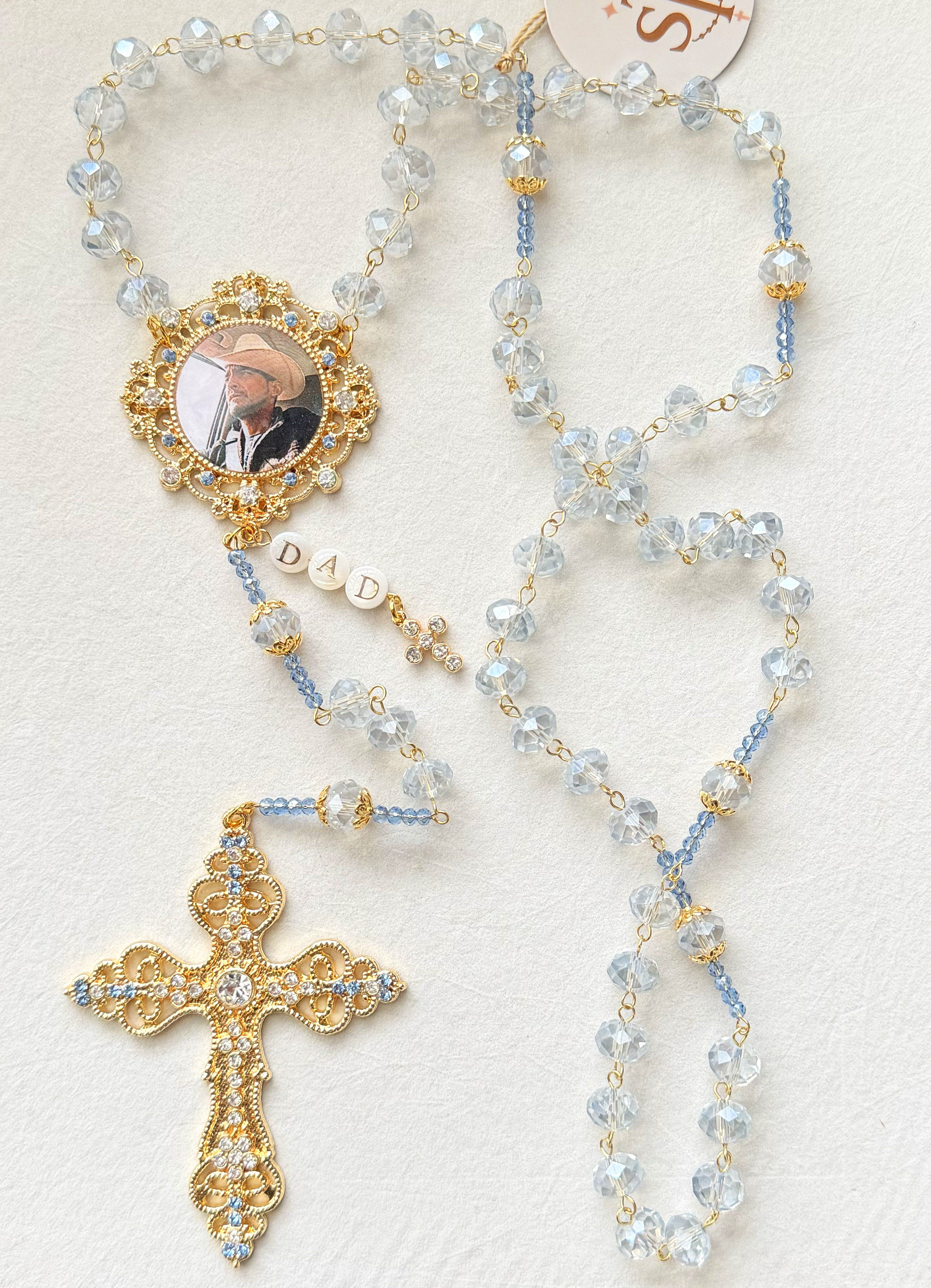 A rosary necklace with clear and blue beads, a gold crucifix, and a religious medallion with a picture of a man wearing a wide-brimmed hat.