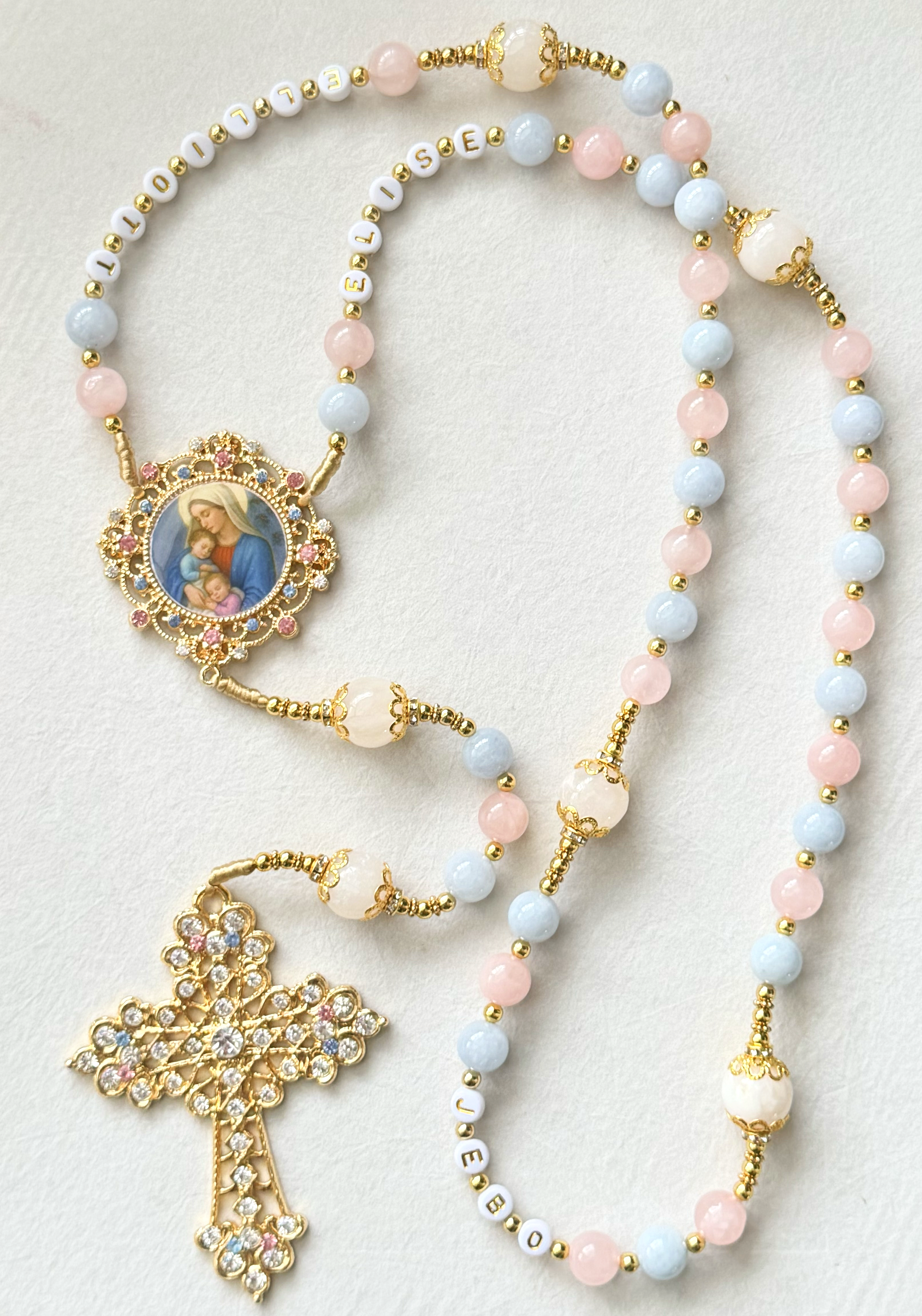 Religion-themed jewelry including rosary beads with pastel pink and blue beads, a pendant with an image of the Virgin Mary holding baby Jesus, and decorative gold and rhinestone elements.