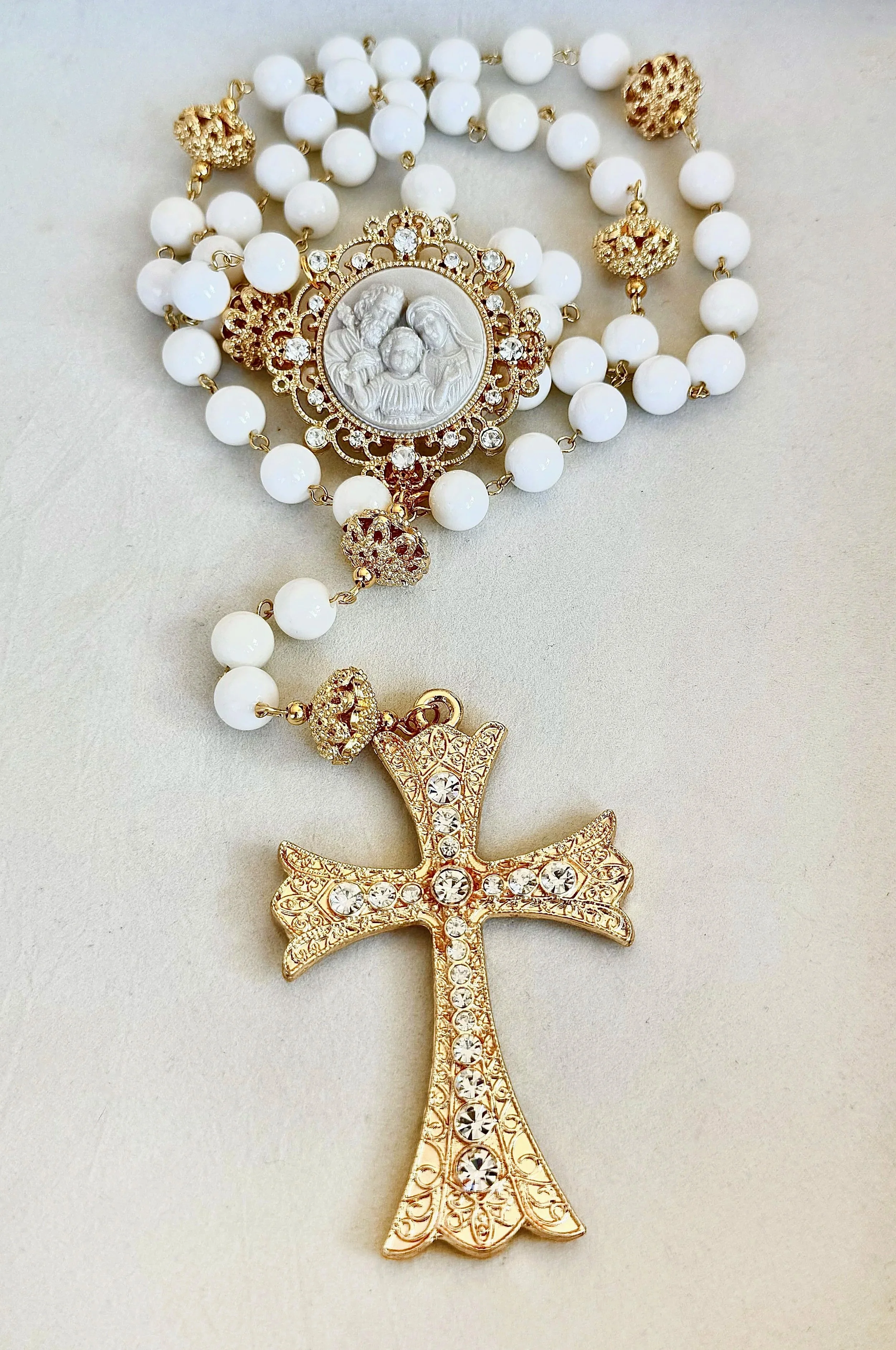 A gold-colored cross pendant adorned with clear rhinestones, with a large religious medallion featuring a carved depiction of the Holy Family surrounded by a decorative border, attached to a beaded necklace with white beads and gold-colored decorativ