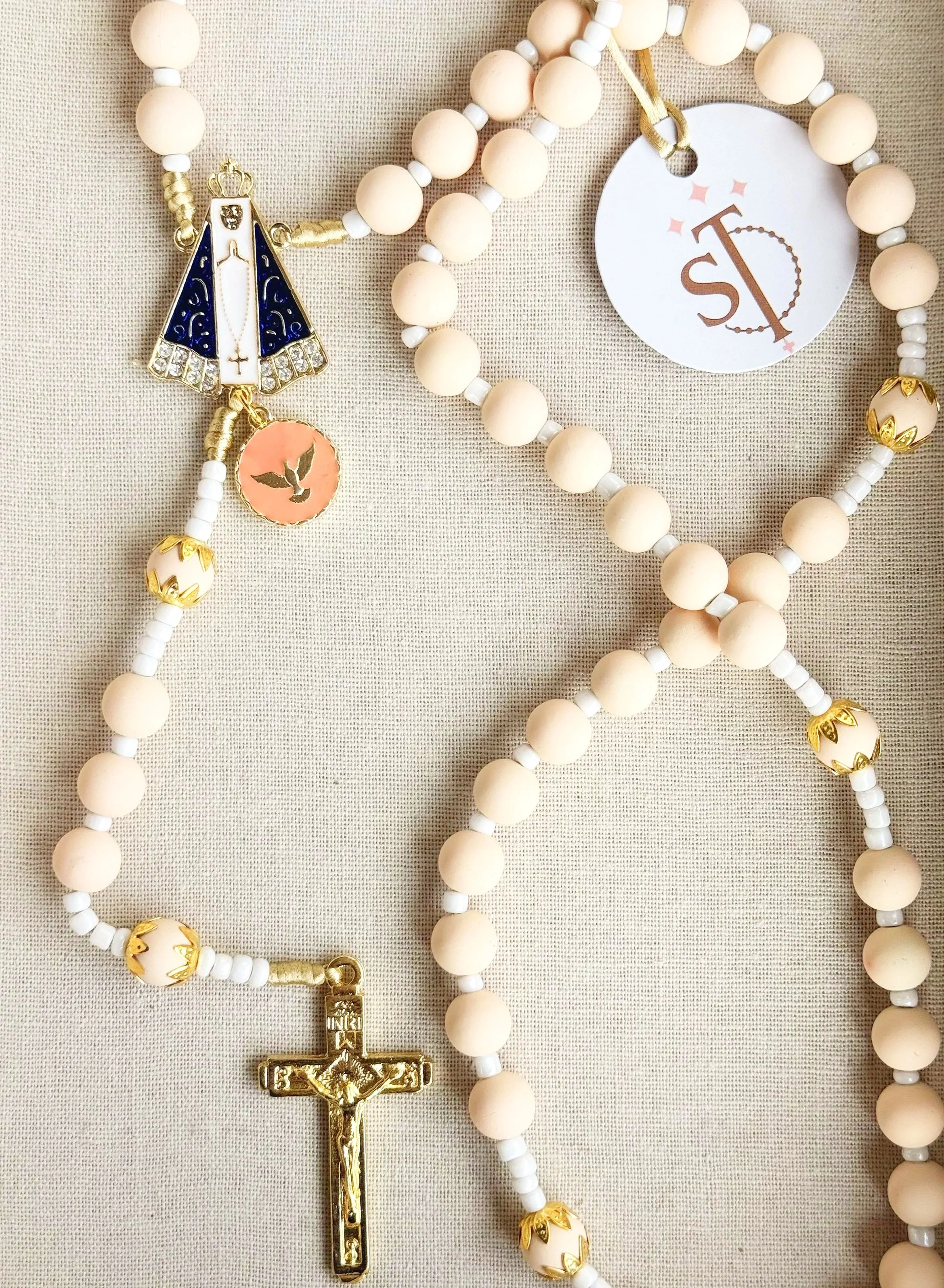 A set of religious rosary beads with cream-colored round beads, gold and white accents, and a gold crucifix at the bottom. The rosary includes a medal with a dove symbol and a small lighthouse charm.