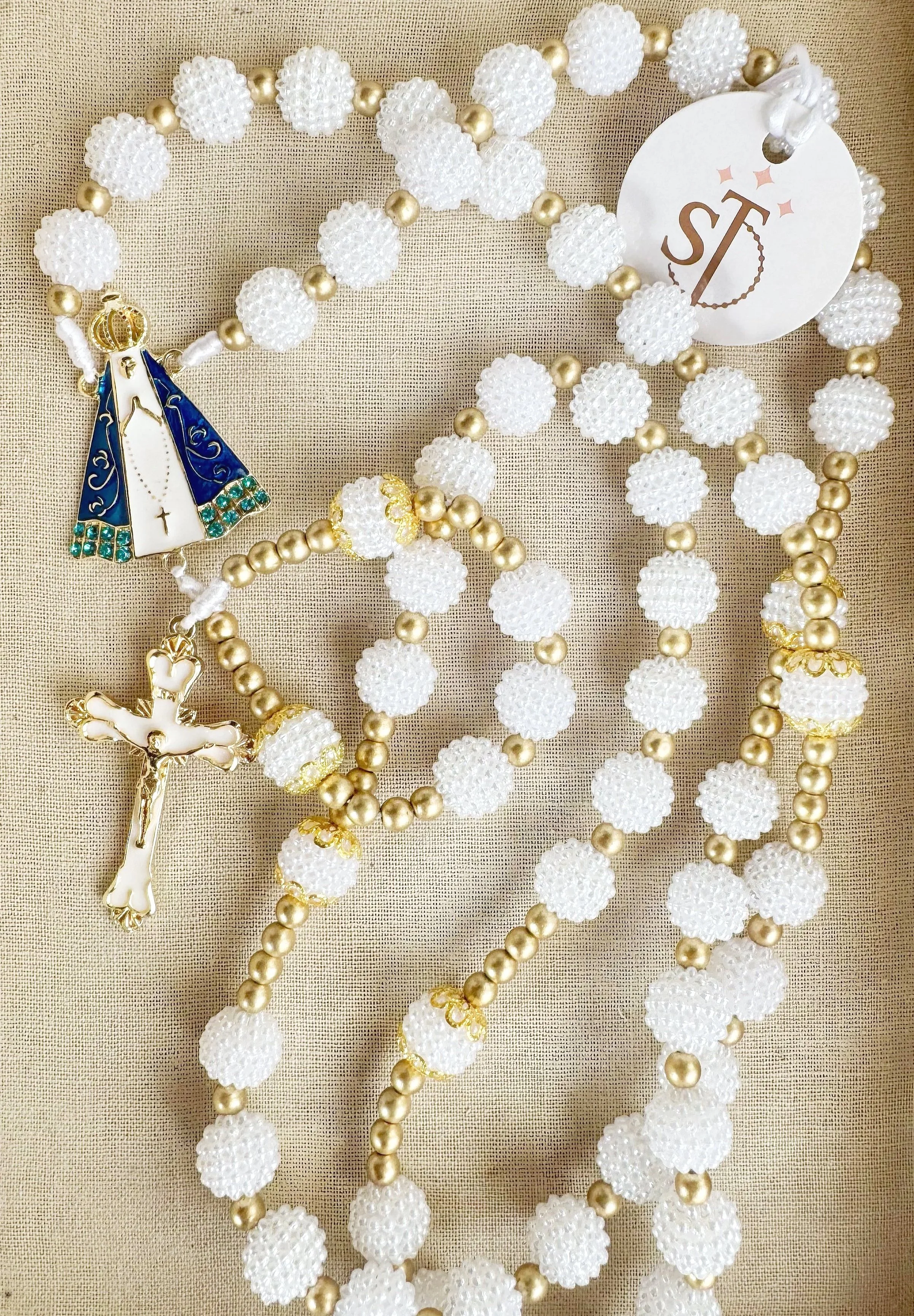 A collection of religious and decorative necklaces on a beige fabric background, including a rosary with white beads and gold accents, a necklace with white and gold beads, and a necklace featuring a religious pendant with a figure of Jesus.