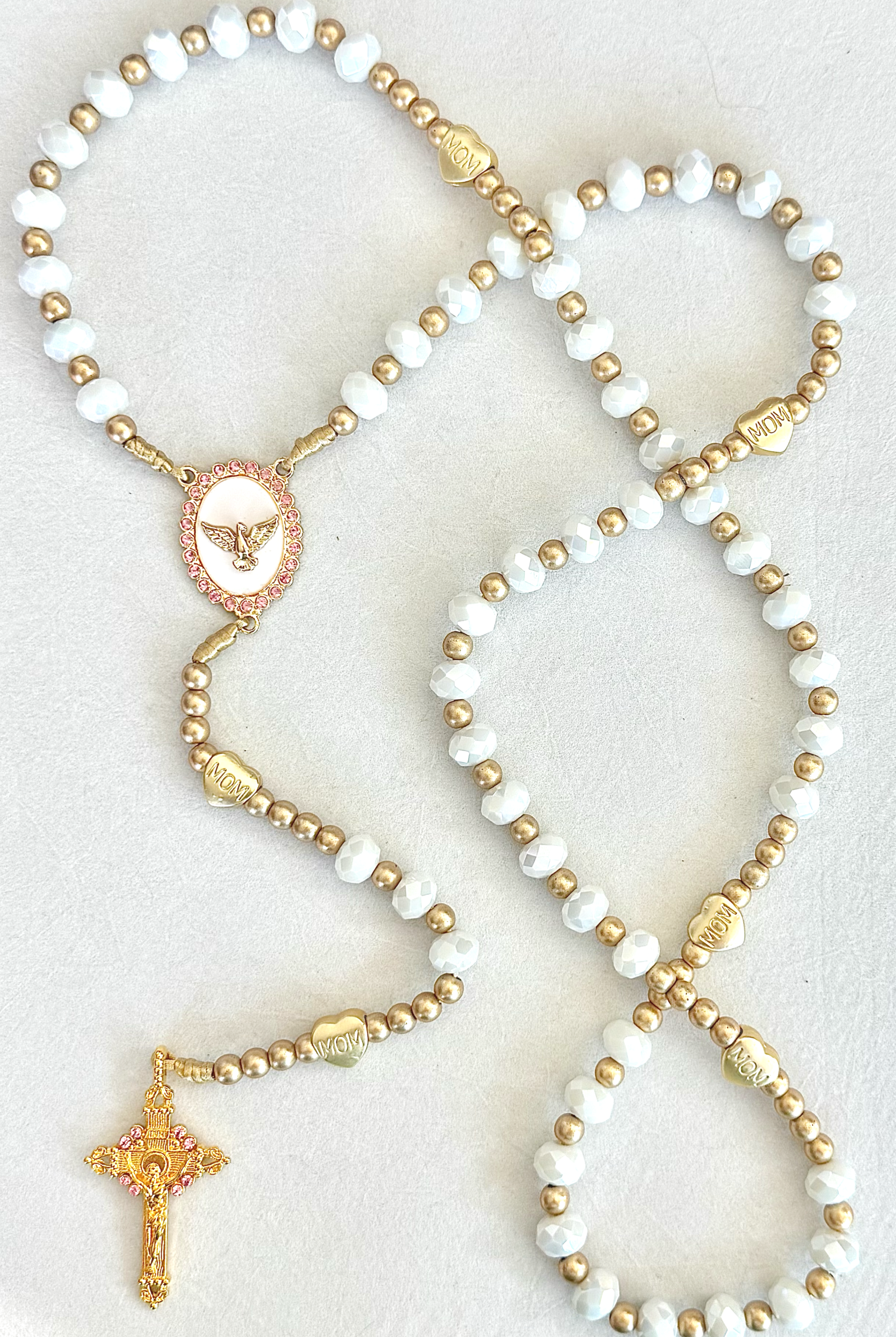 Beaded necklace with white and gold beads, a gold heart with 'MOM' engraved, and an ornate gold pendant featuring an angel figure, on a white background.