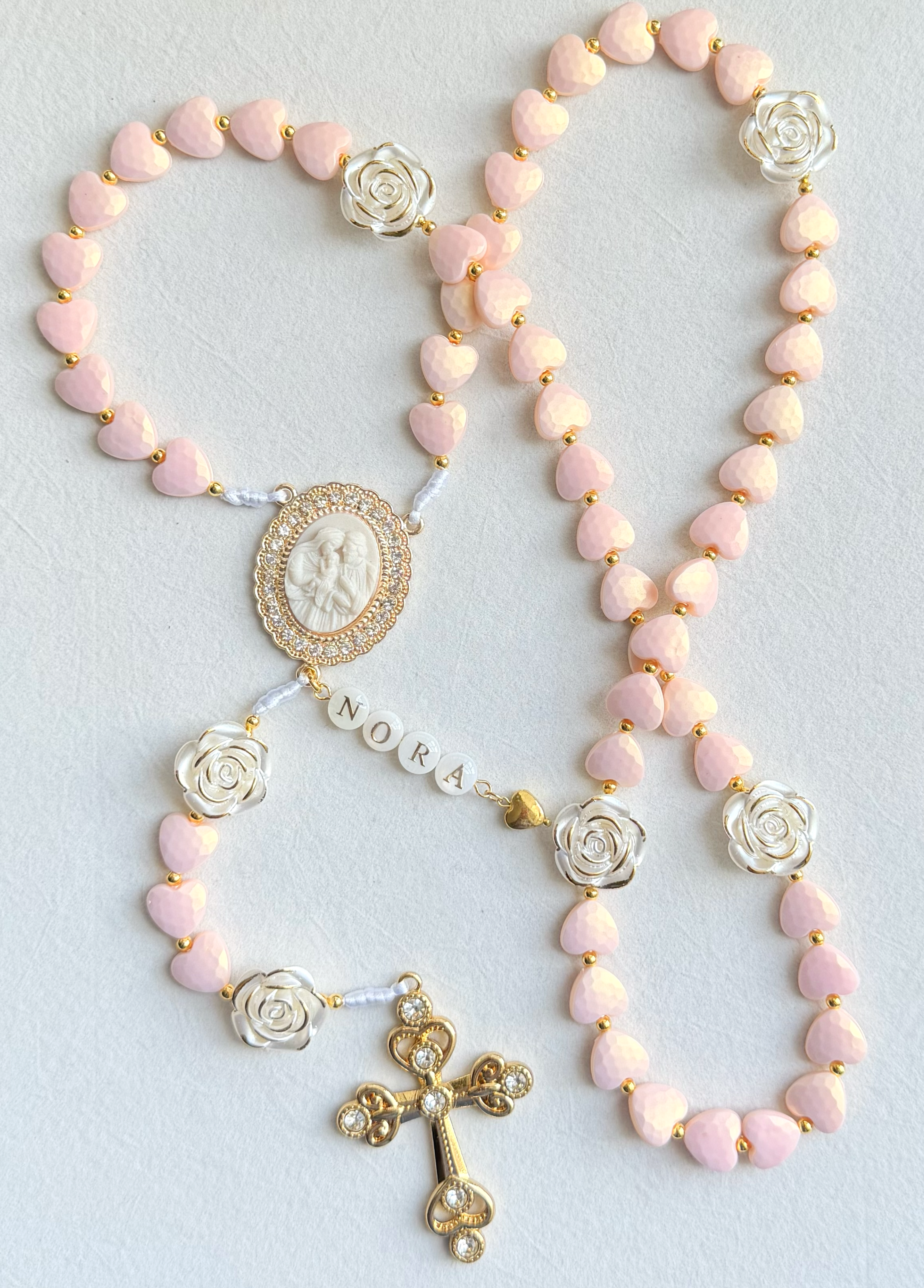 A rosary with pink heart-shaped beads, silver rose beads, a white religious cameo, and a gold cross pendant with stones.