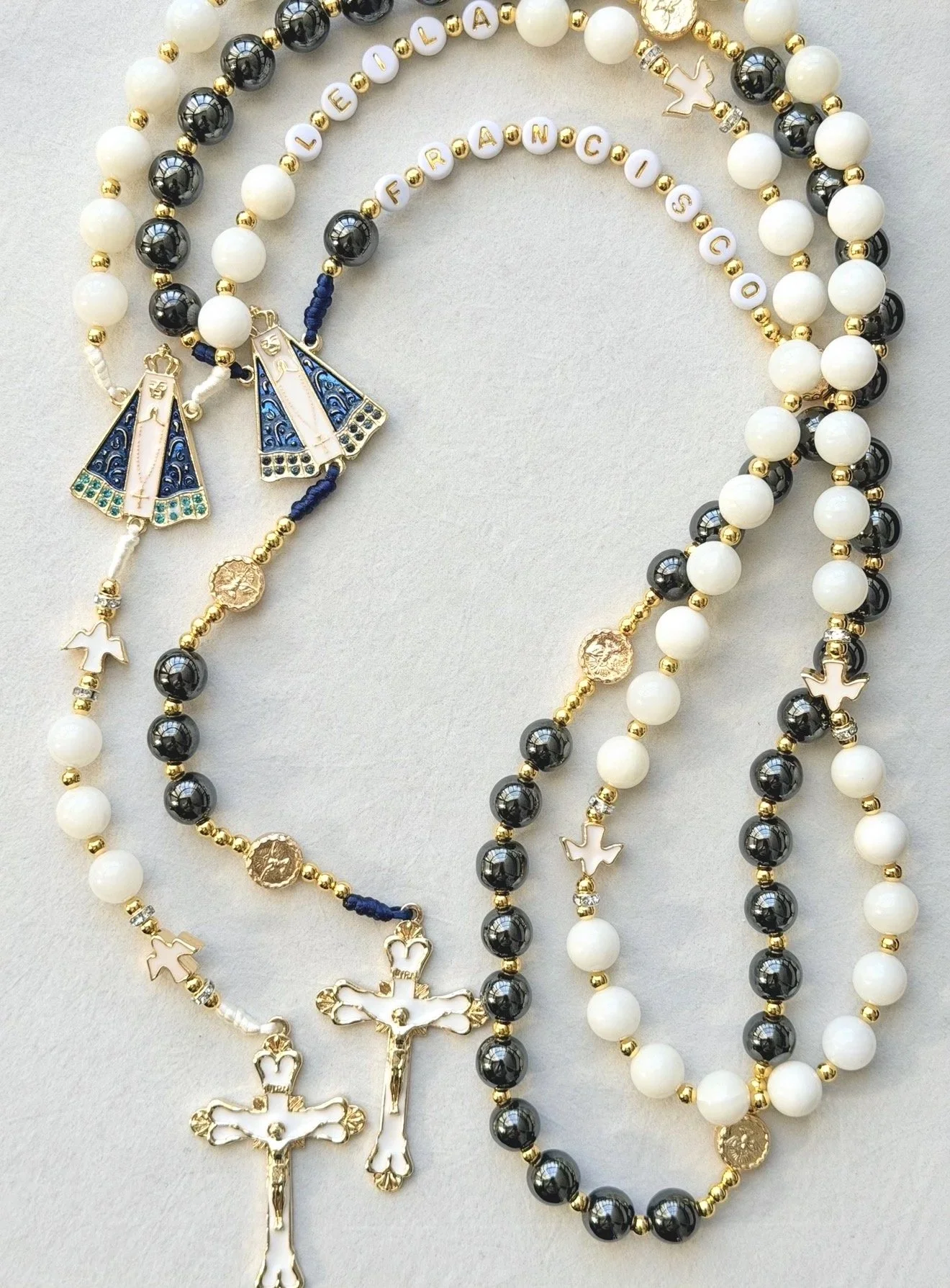 A collection of Catholic rosary necklaces with white, black, and gold beads, featuring medals, crucifixes, and statues of the Virgin Mary.