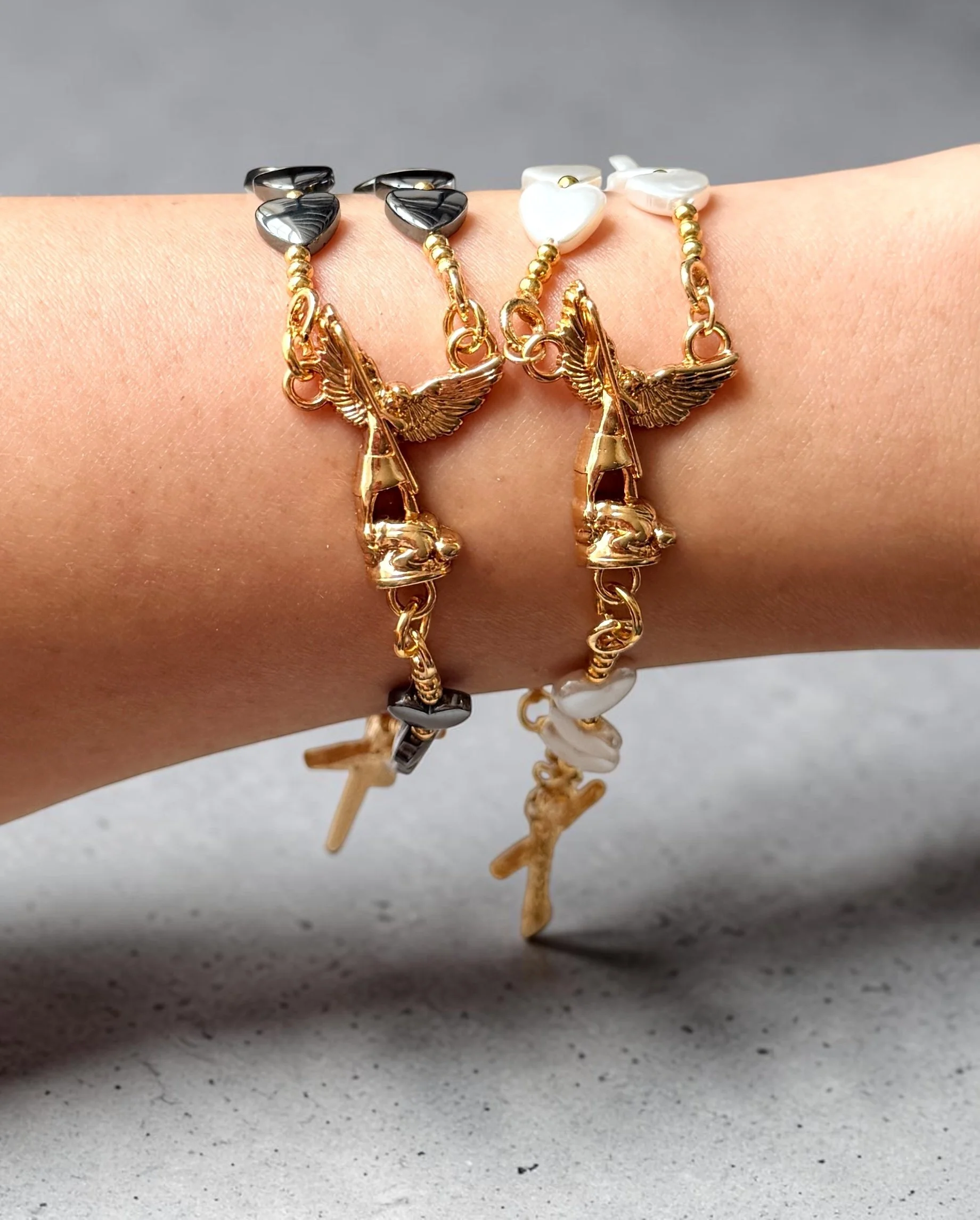 Multiple bracelets with heart-shaped charms and gold chain links, featuring angel and star pendants, displayed on a person's wrist against a neutral background.