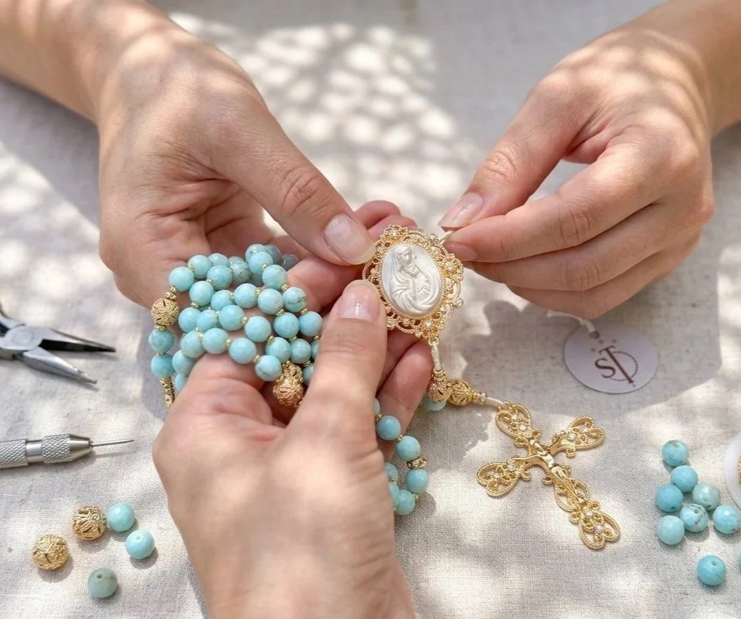 Hands assembling gold and pearl necklaces and religious medals on a crafting table.