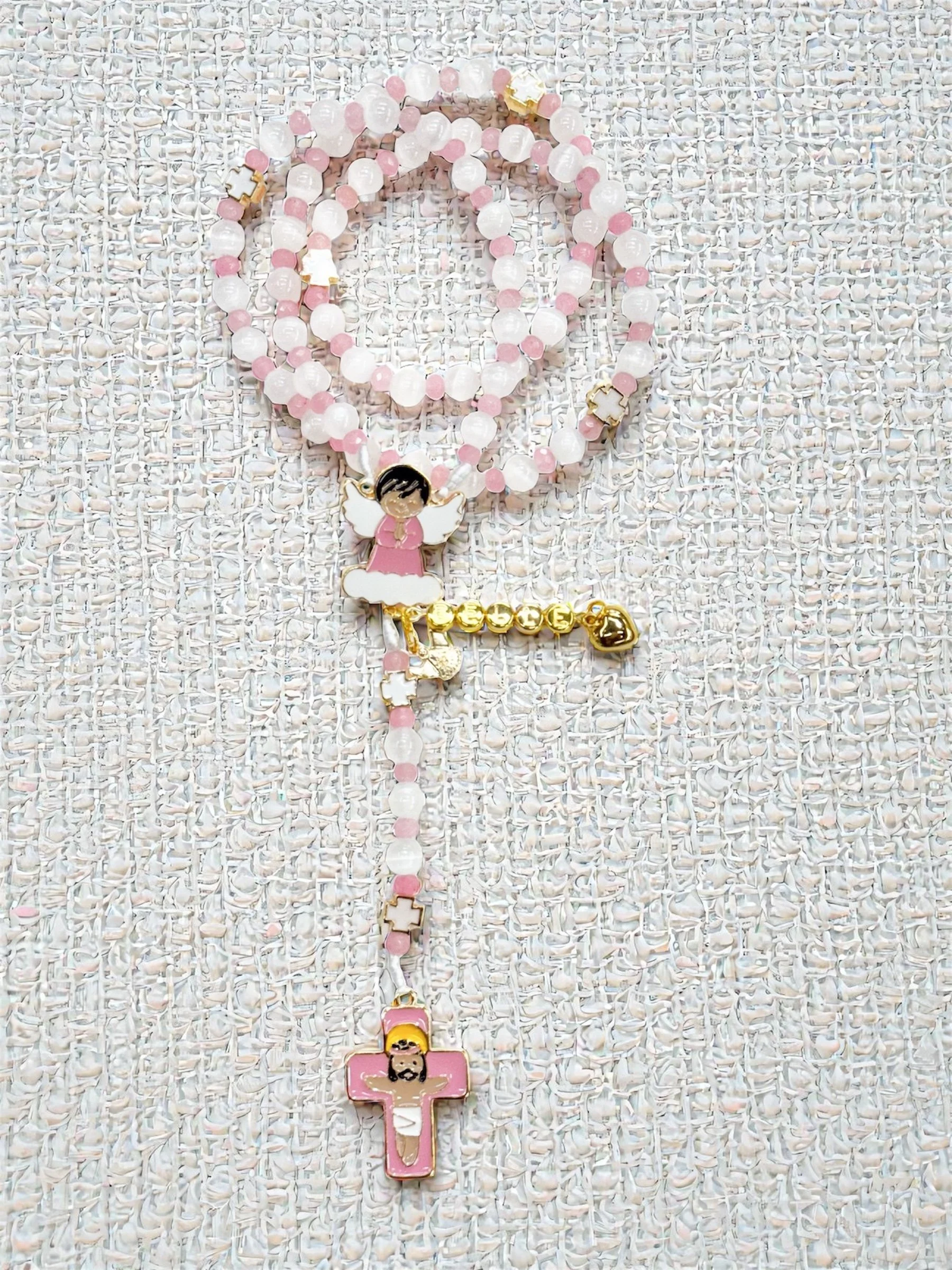 A rosary with pink and white beads, featuring a charm of an angel with black hair, pink dress, and white wings, and a crucifix with a depiction of Jesus Christ, both attached to a white and pink beaded chain, laid on a textured white background.