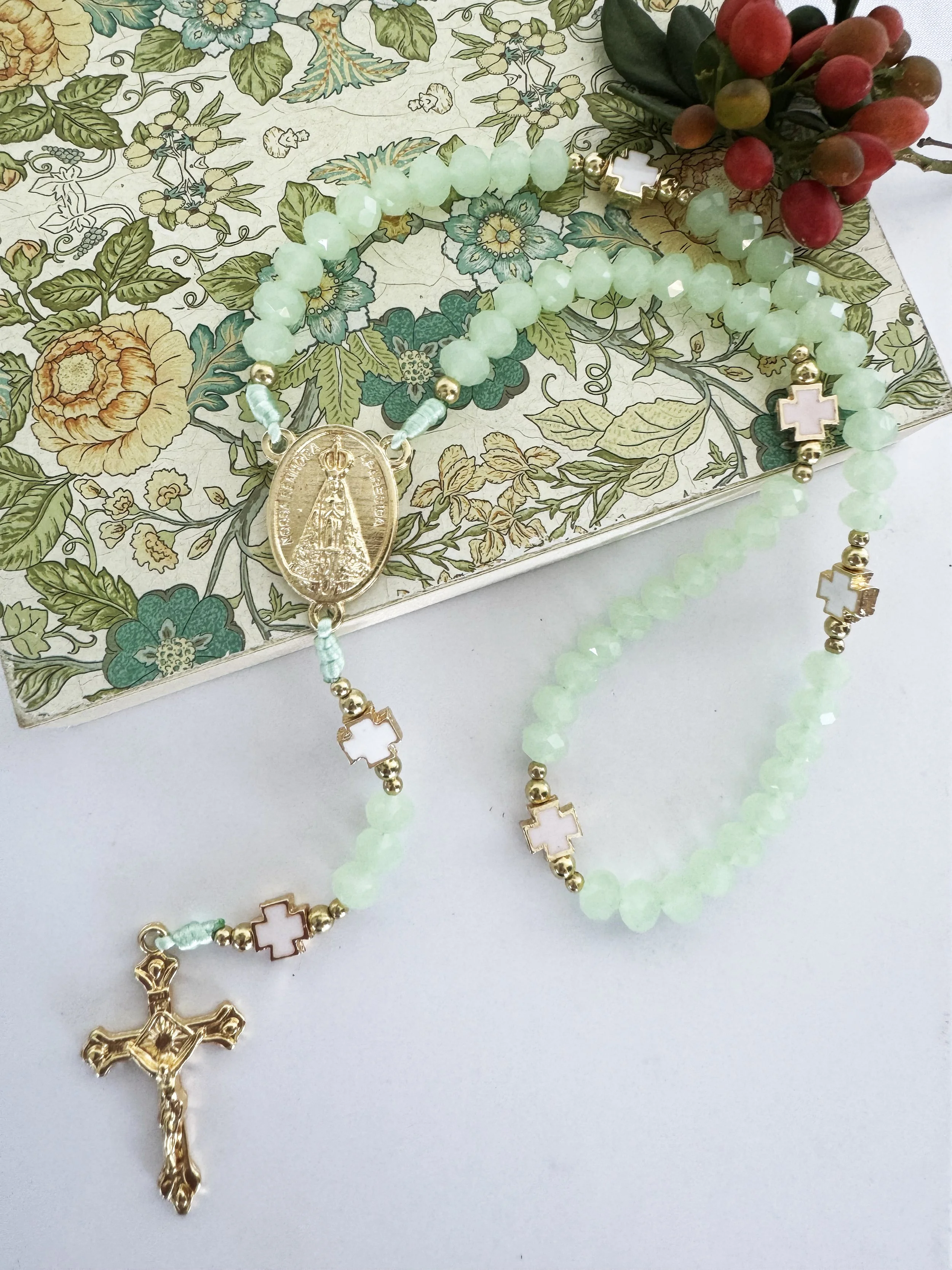 Two rosary necklaces with gold, green beads, and white cross pendants are draped over a floral illustration background.