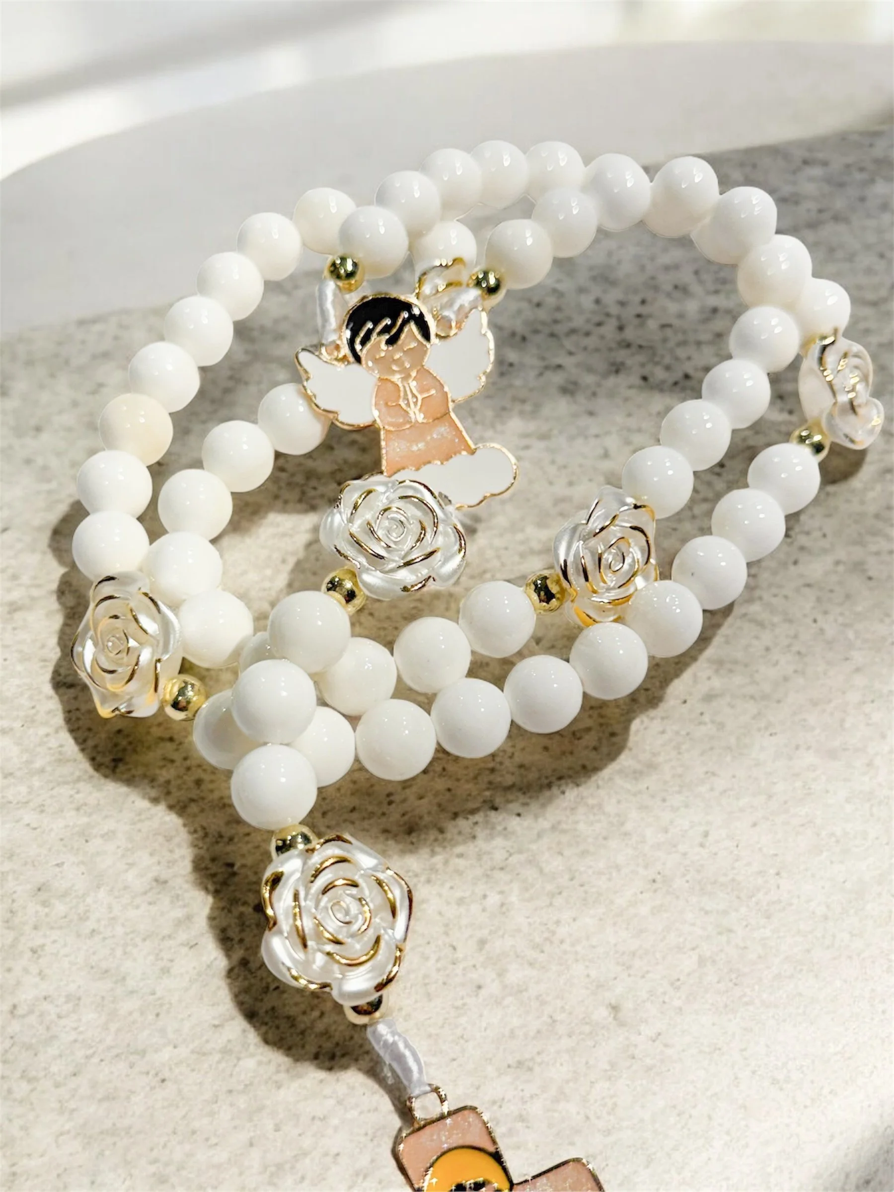 Close-up of an angel-shaped enamel pin with pink dress, black hair, and white wings, surrounded by white pearl beads and gold floral accents on a gray surface.