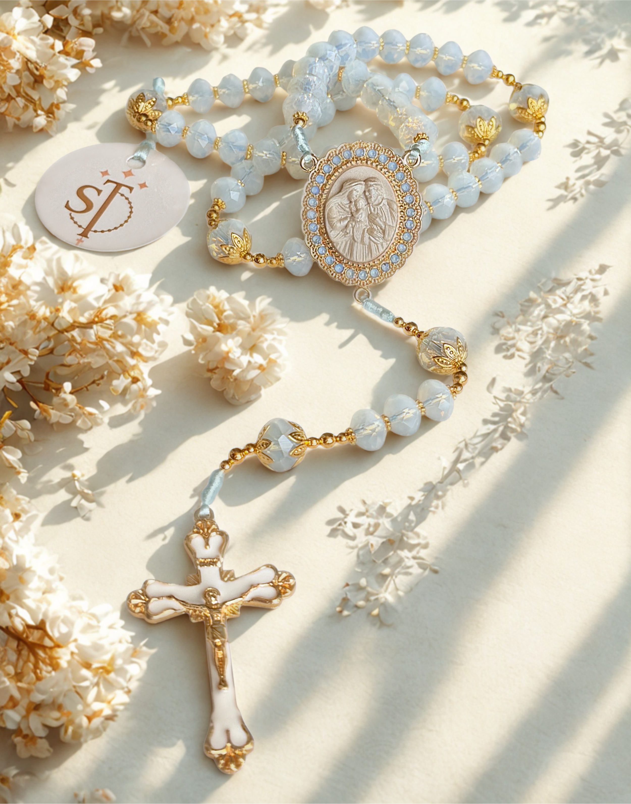A religious rosary with white and gold beads, featuring a medallion with an image of the Virgin Mary, laid on a light surface surrounded by white flowers.