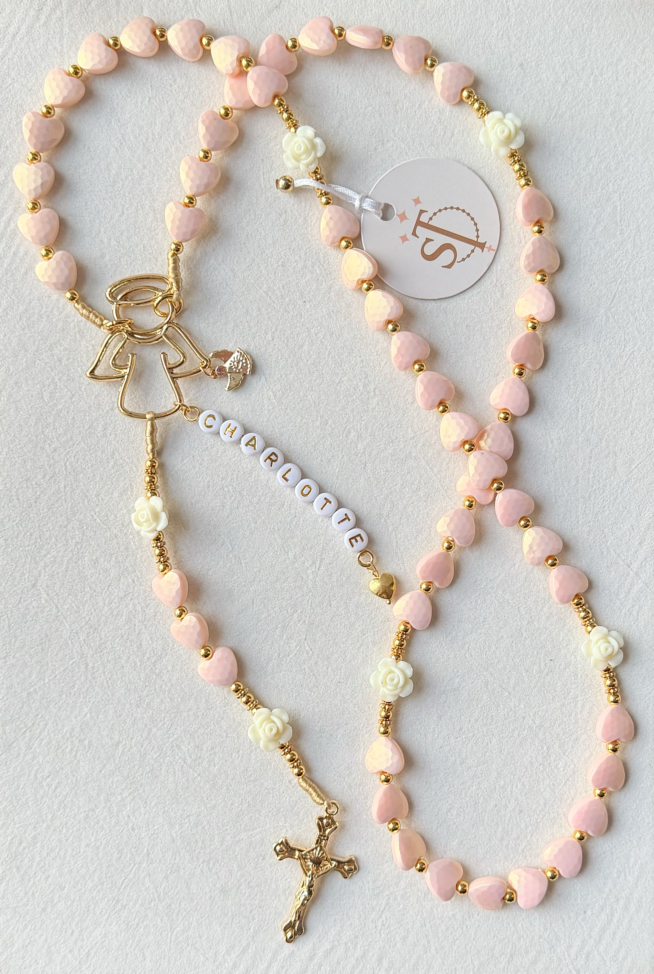 A pink and gold beaded rosary with small heart-shaped pink beads, white rose beads, and a gold crucifix at the bottom.