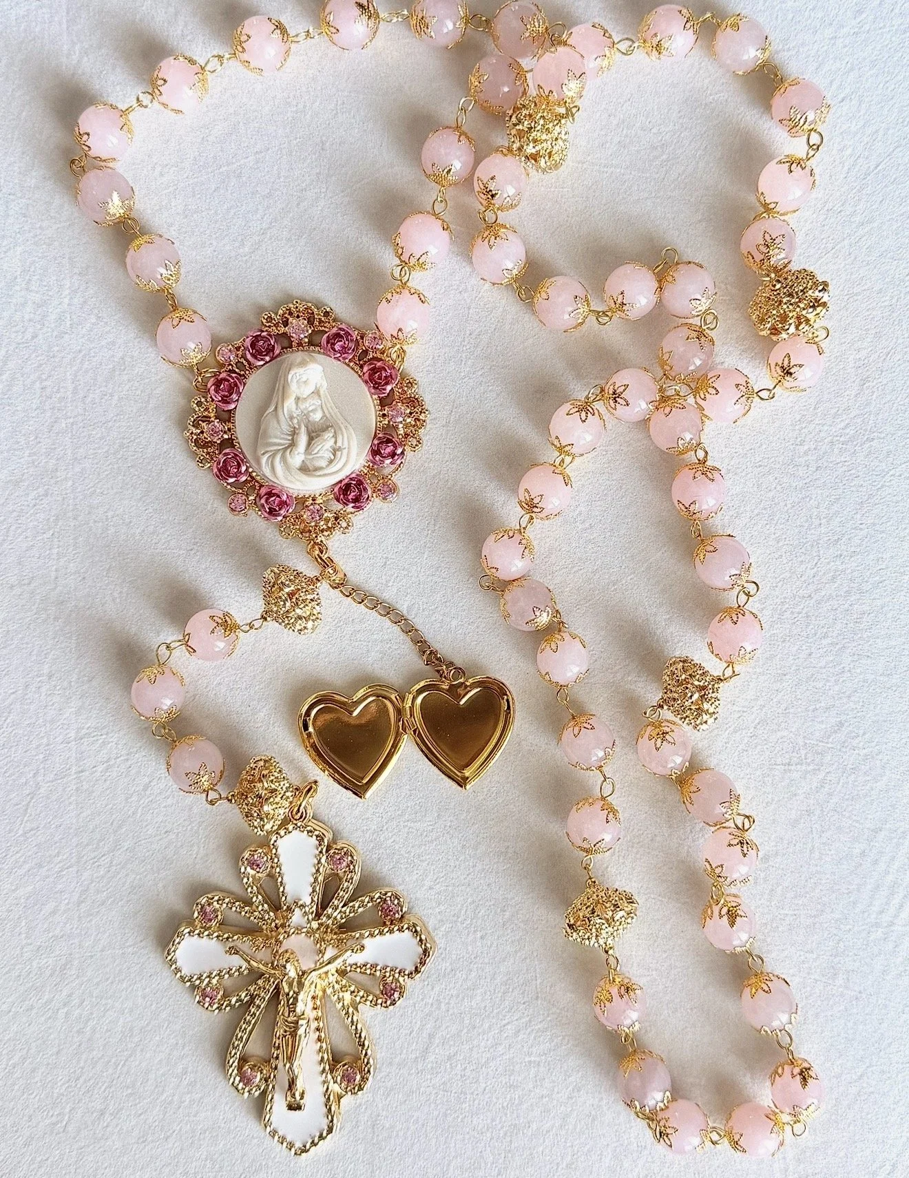 A set of pink rosary beads with gold accents, featuring a religious medallion depicting Mary and Baby Jesus, along with gold heart-shaped and butterfly-shaped accessories, arranged on a white surface.