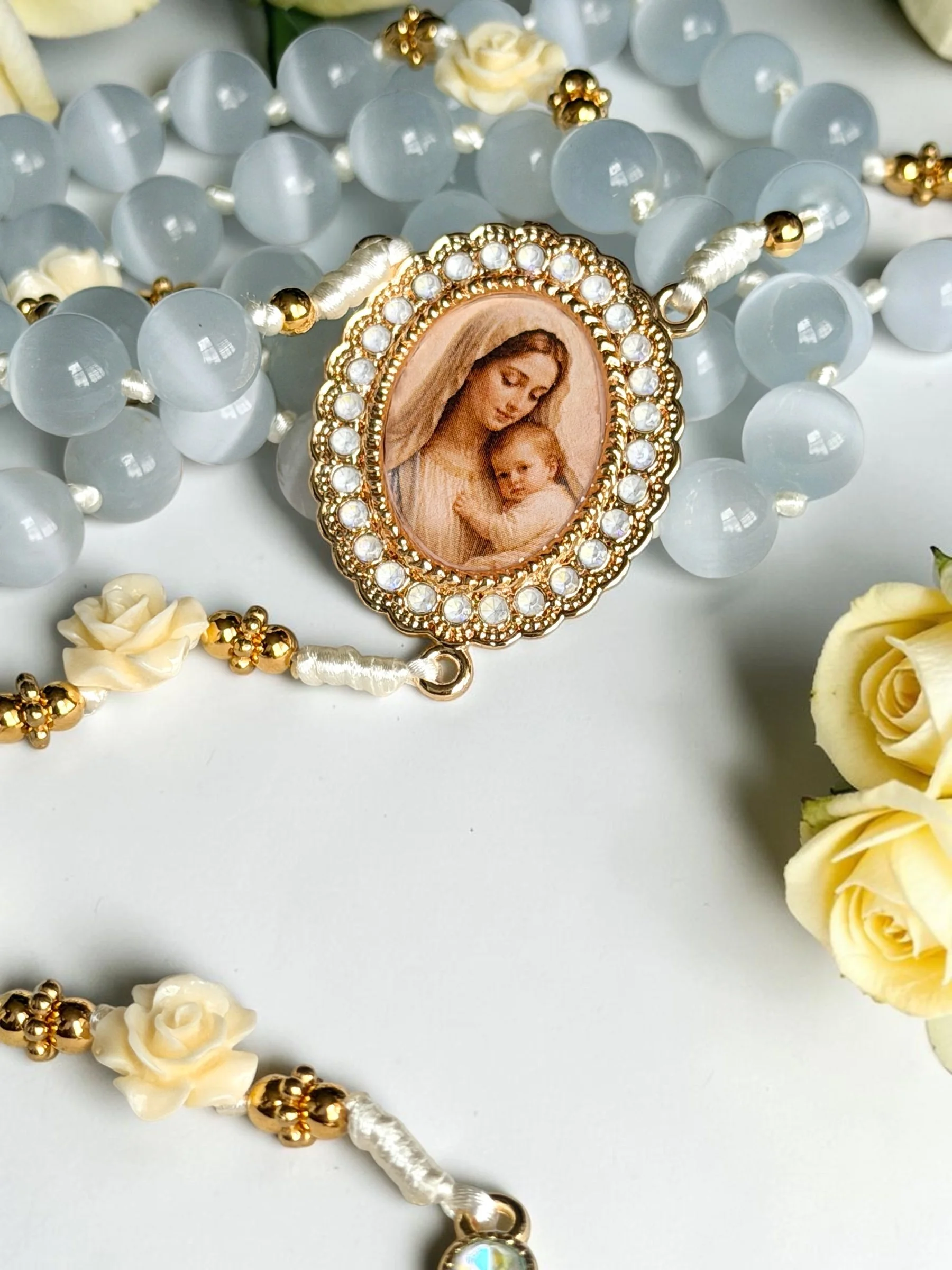 A rosary with a pendant featuring an image of the Virgin Mary and baby Jesus, surrounded by white and gold beads, laid on a white surface with yellow roses nearby.