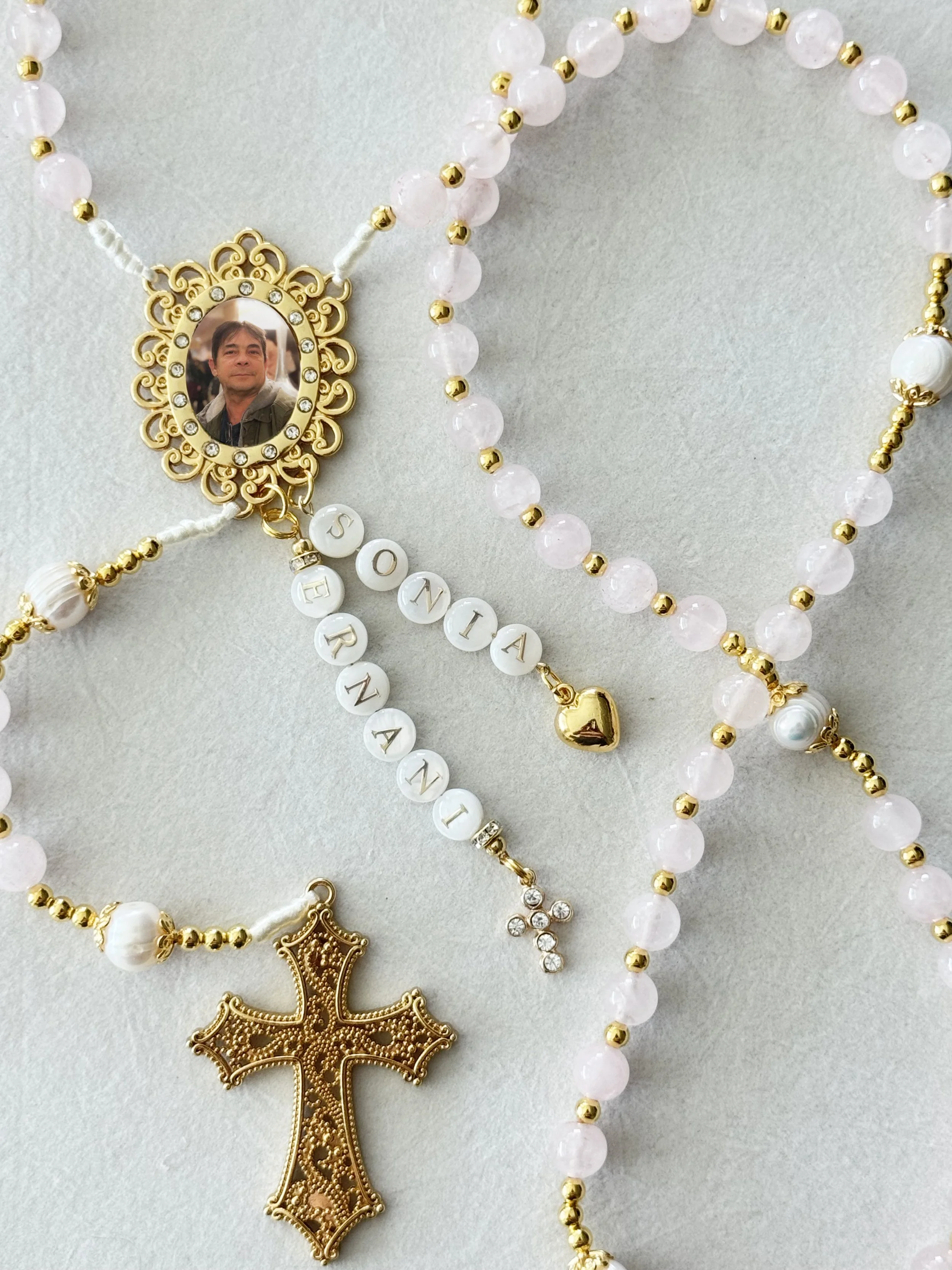 A collection of jewelry including necklaces with beads, gold accents, and pendants featuring a man's photo, heart charms, and decorative elements on a light background.