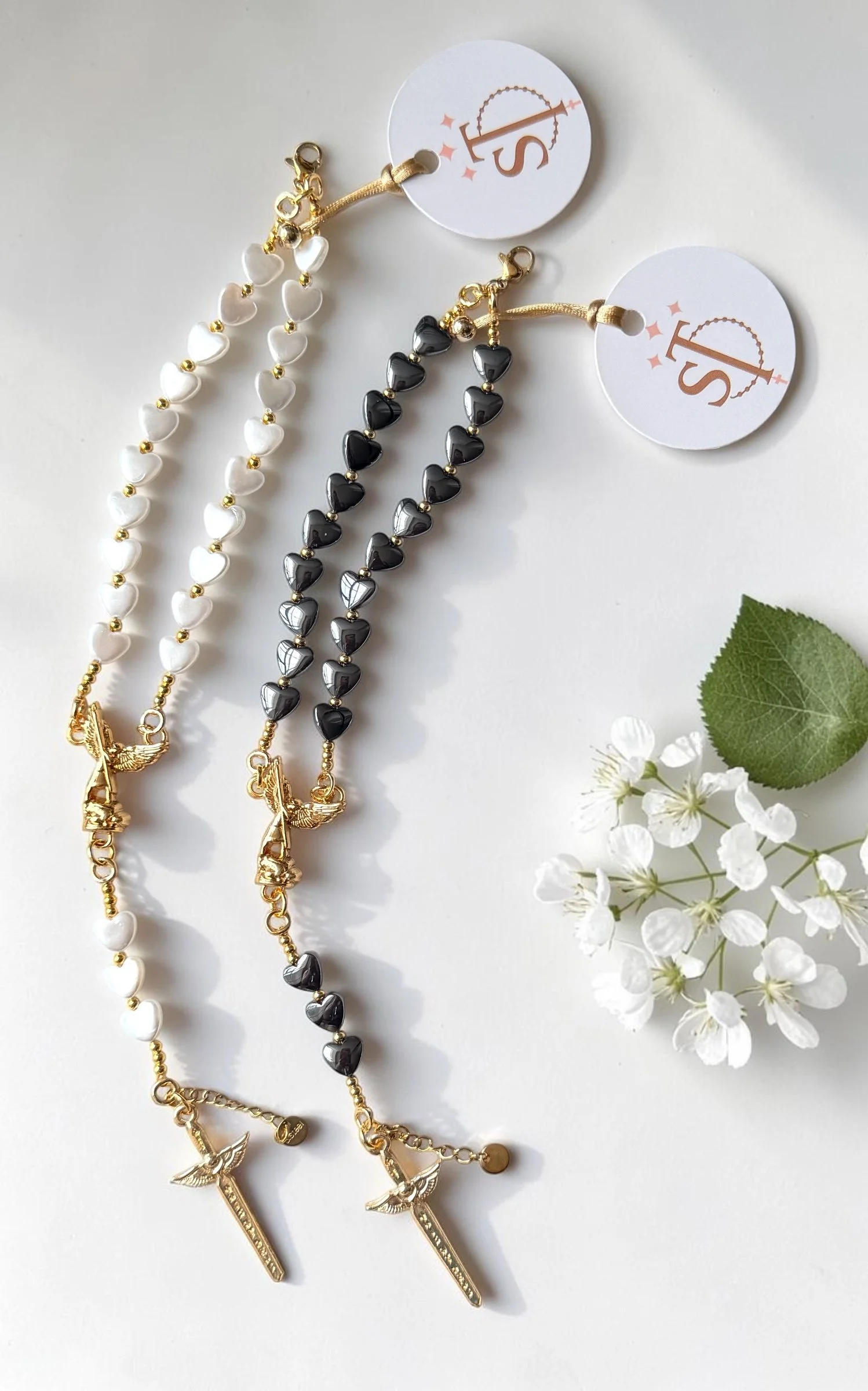 Two necklaces, one with white heart-shaped beads and the other with black heart-shaped beads, both with gold charms including a sword, a winged emblem, and a crown.