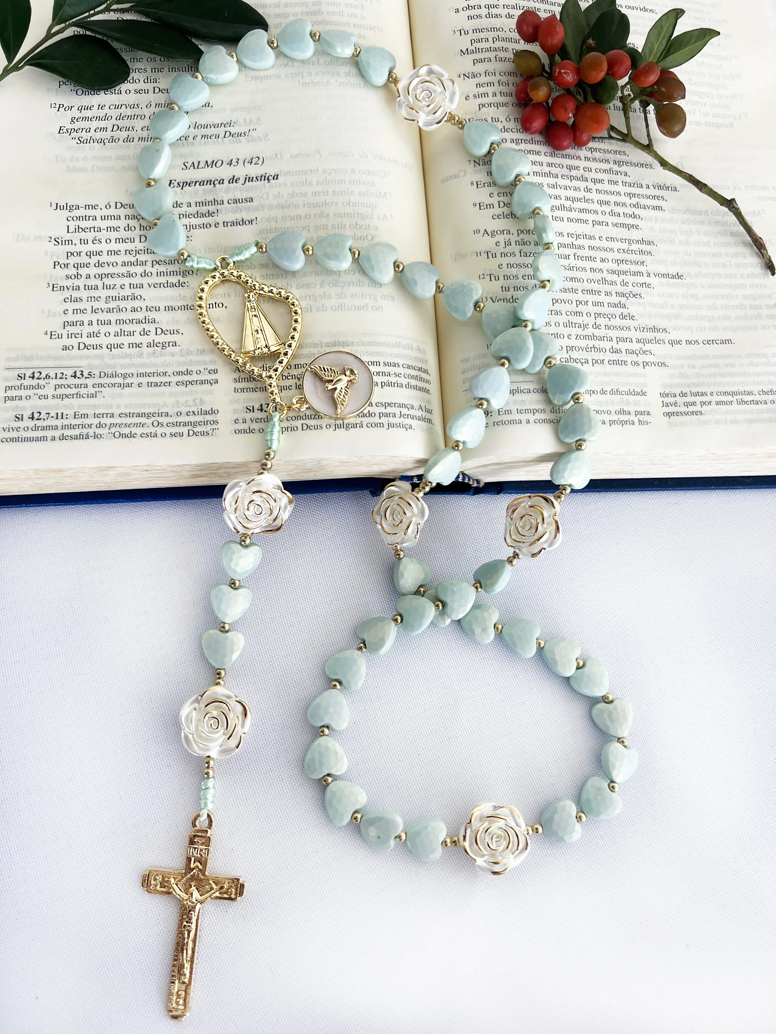 Light blue heart-shaped beads and white rose-shaped beads on a gold chain with a golden crucifix and a small golden dove pendant, resting on an open Bible with red and black text, accompanied by green leaves and red berries.