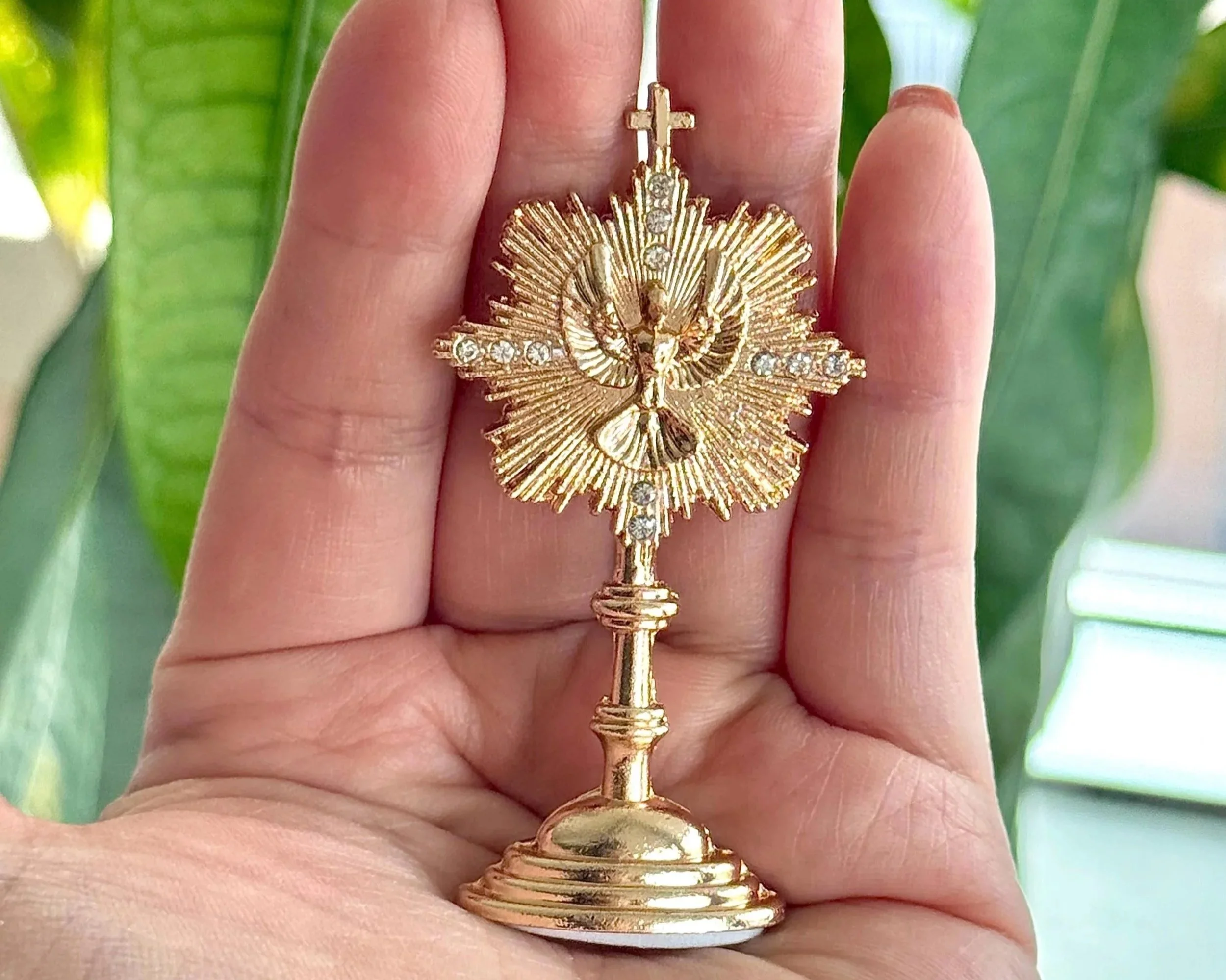 Gold religious pendant featuring a crucifix and a radiant figure with a crown of rays, adorned with small gems, held between fingers with a green background.