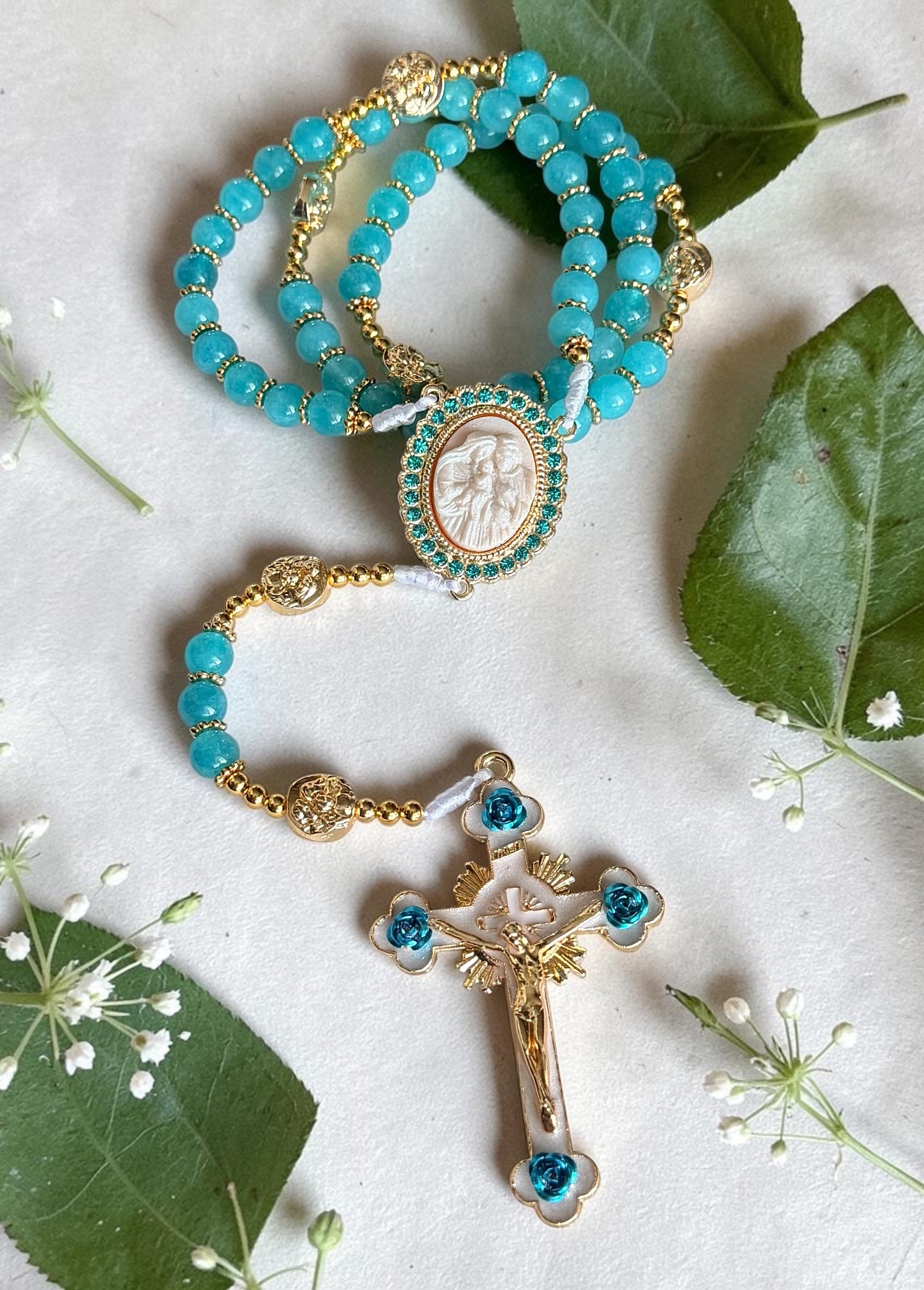 A gold and turquoise rosary with blue beads and a religious pendant featuring a carved figure, laid on white fabric with green leaves and small white flowers around it.