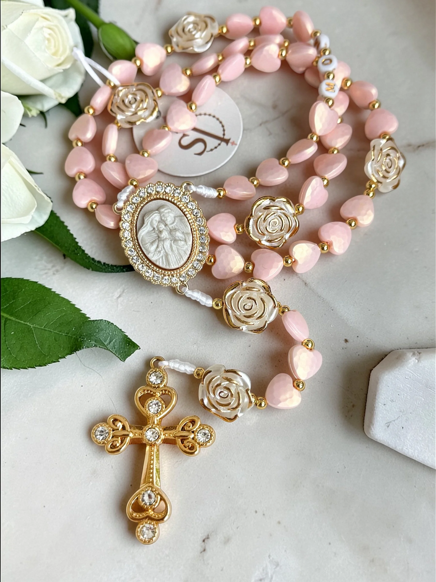 Close-up of a pink beaded rosary with pearl and gold flower accents, a cameo medallion of a religious figure, a gold crucifix with rhinestones, and white roses with green leaves on a white surface.