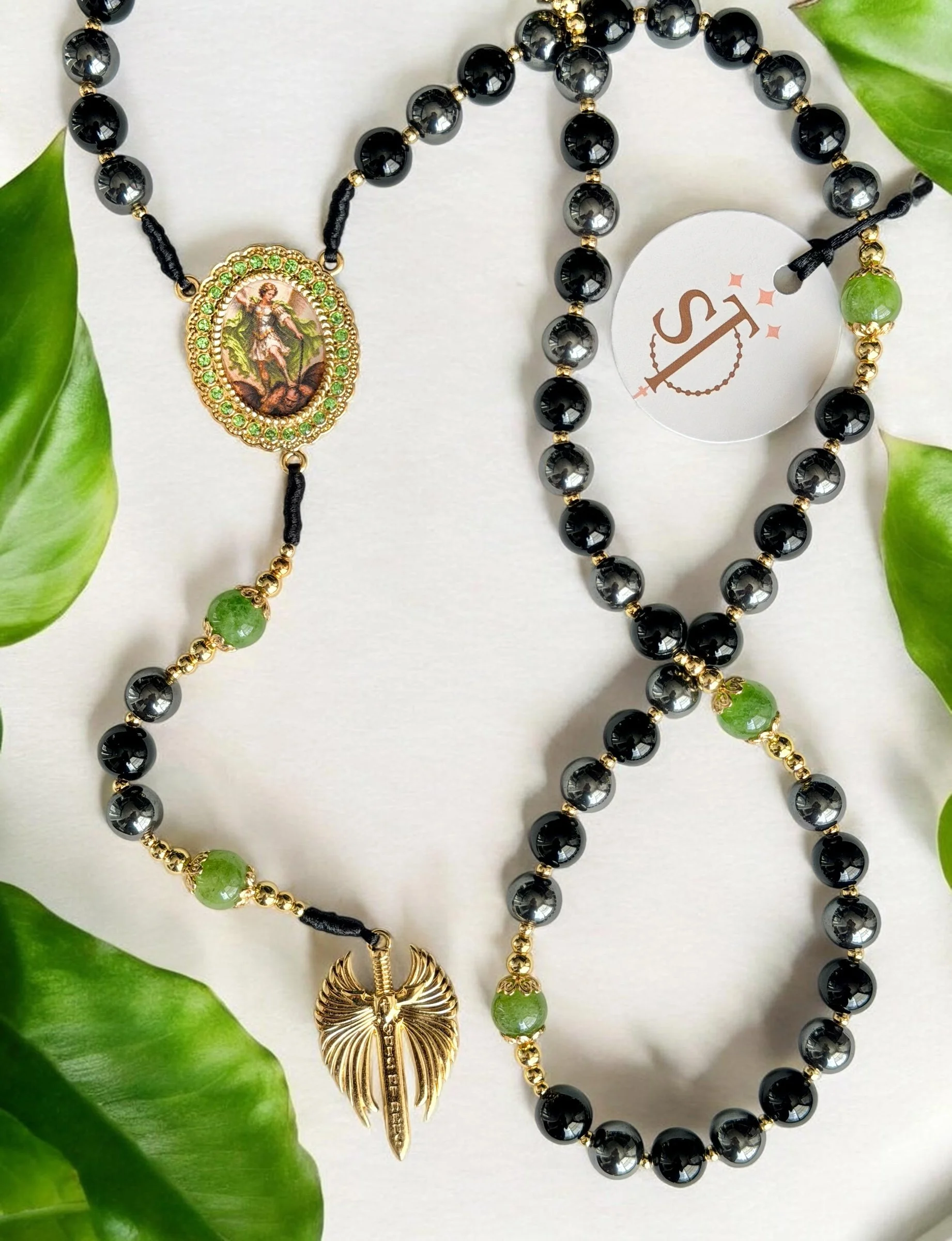 A gold and black beaded necklace with a large gold and green pendant, a second multi-colored beaded necklace with a gold shell charm, and a gold and black beaded bracelet, all resting on a white surface with green leaves.