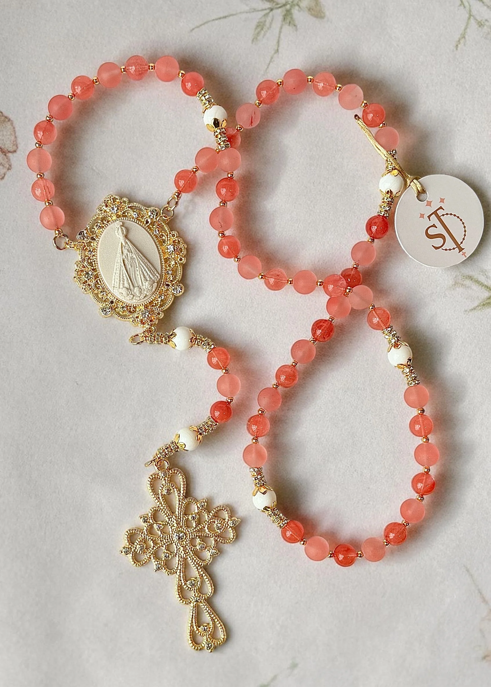 Pink beaded necklace with gold accents and a religious figurine pendant, gold ornate snowflake-shaped pendant, and a white circular tag.