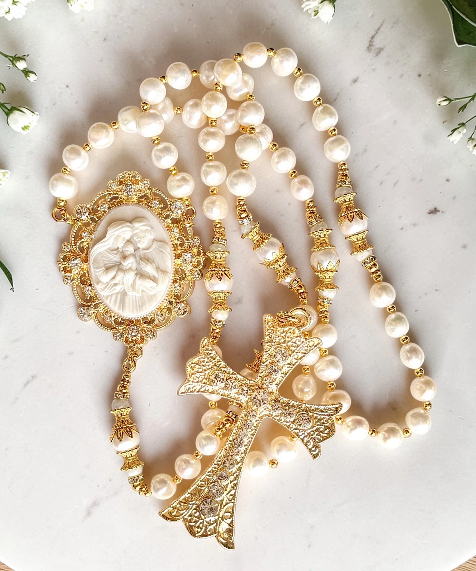 Close-up of pearl and gold jewelry, including necklaces, and brooch with a central cameo depicting a bearded man. The jewelry has intricate gold details and sparkling stones, arranged on a white surface. Small white flowers are visible at the edges.