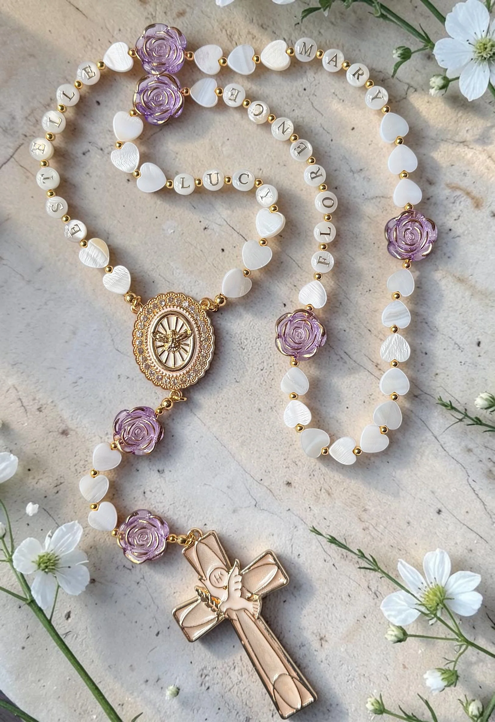 A collection of jewelry including a white and gold beaded necklace with purple rose accents, a smaller matching bracelet with beads spelling "LUCKY," a gold oval pendant with a sun and moon design, and a gold angel charm holding a gift box, all on a 