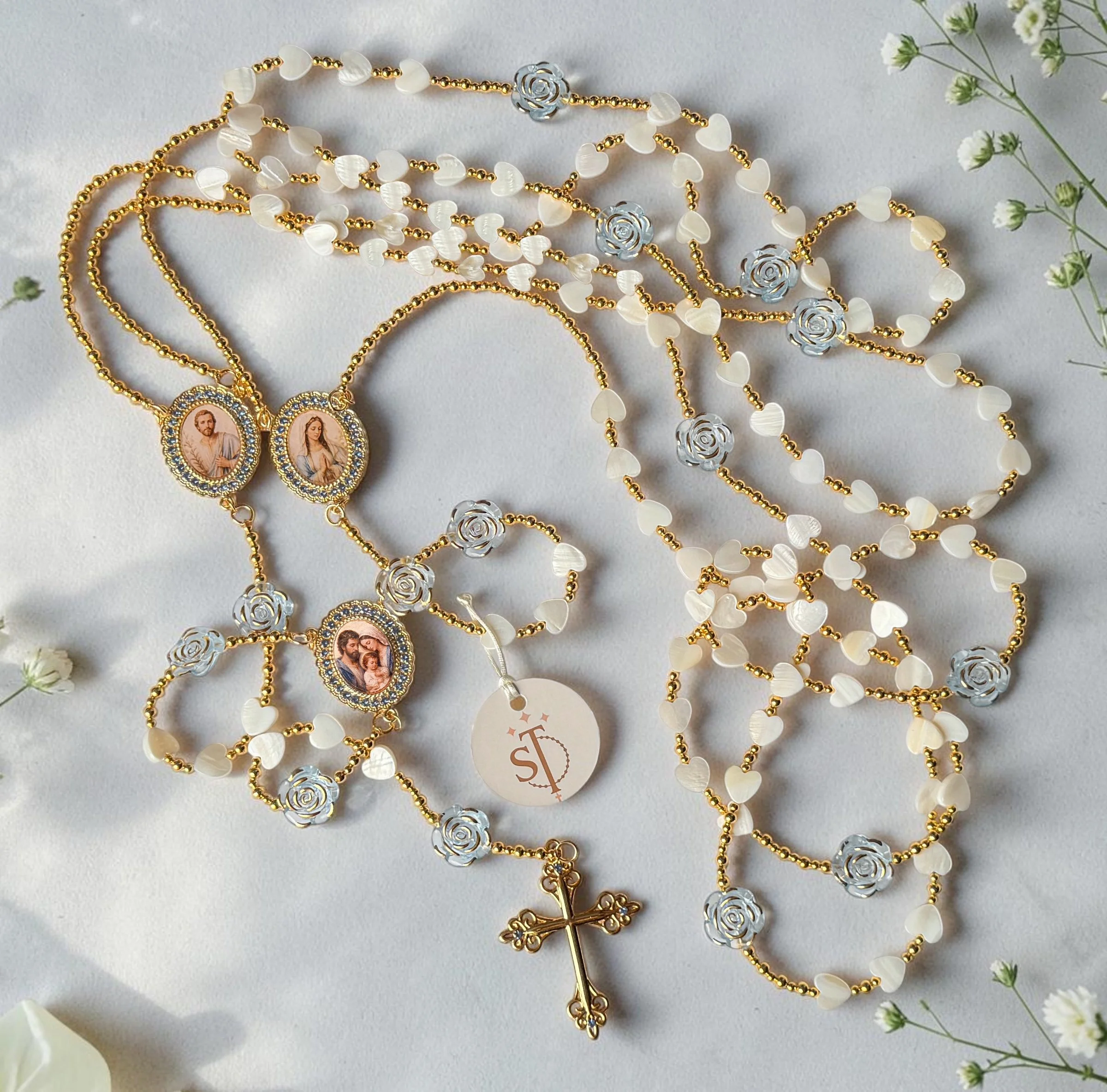 Necklaces with religious icon pendants, gold chains, white heart-shaped beads, silver rose pieces, and gold cross, arranged on a white surface with small white flowers nearby.